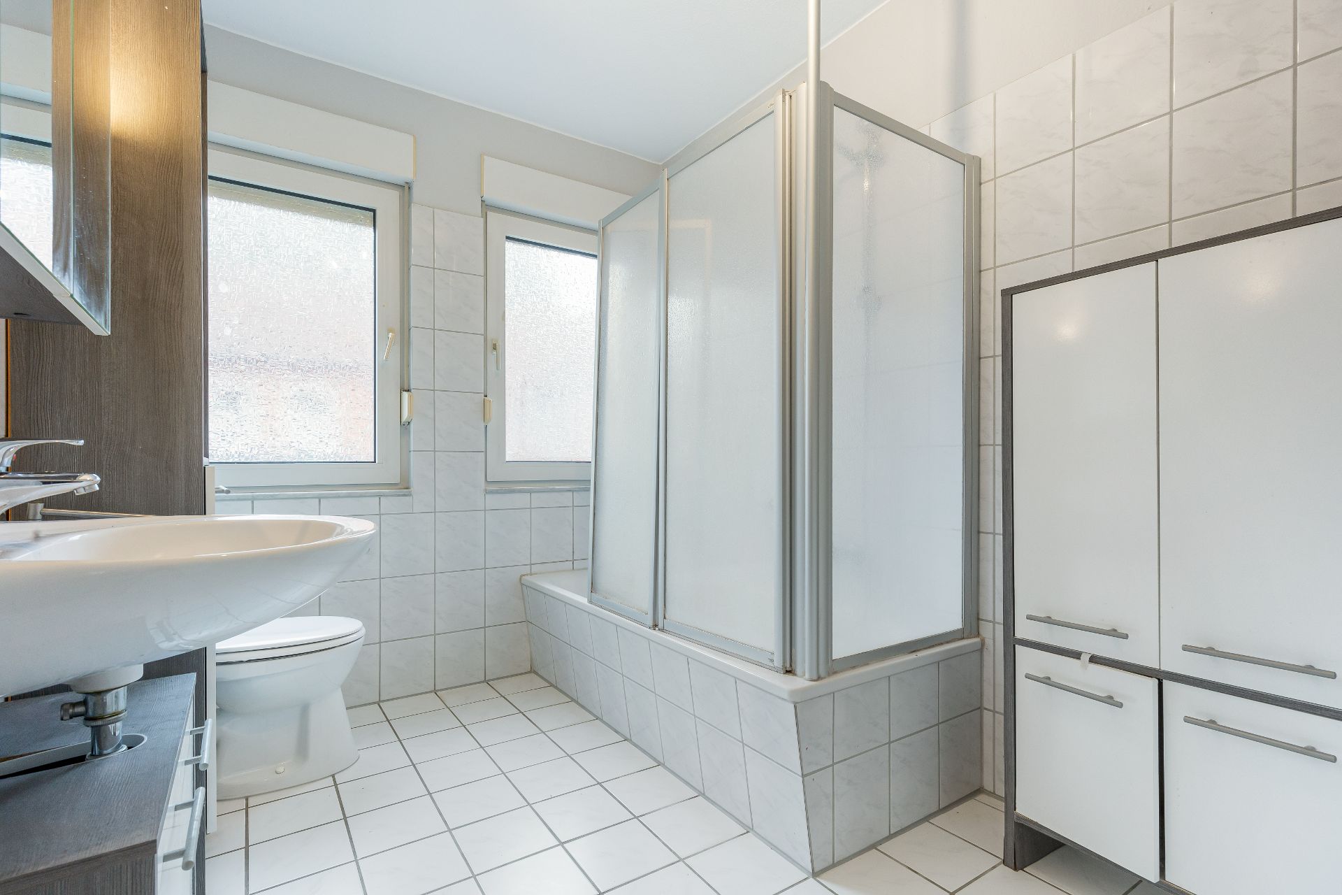 Helles Bad mit Fenster, Dusche in Badewanne, Waschbecken, Toilette und weißem Schrank.