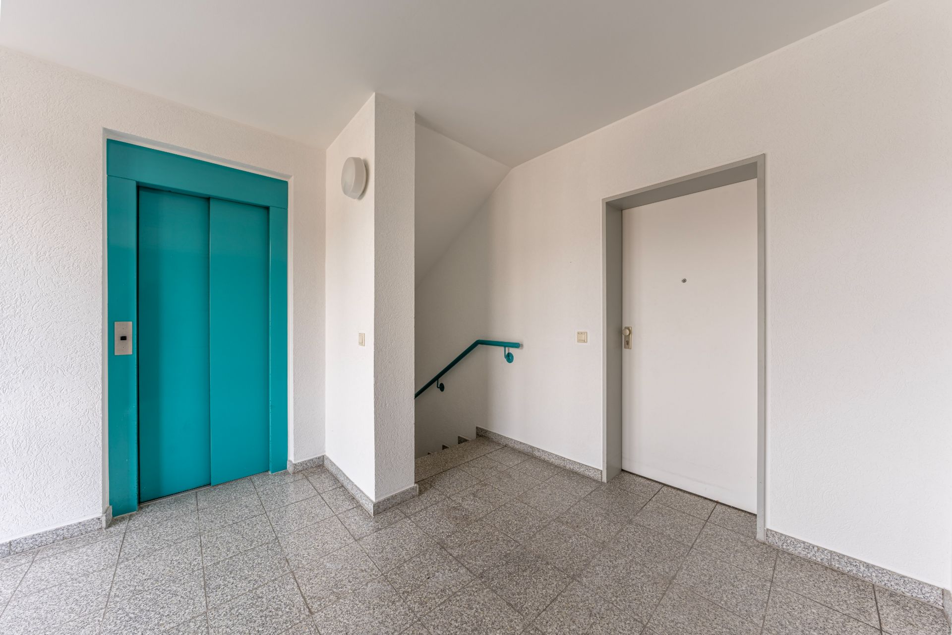 Heller Flur mit türkisfarbenem Aufzug, Treppe und weißer Wohnungstür.