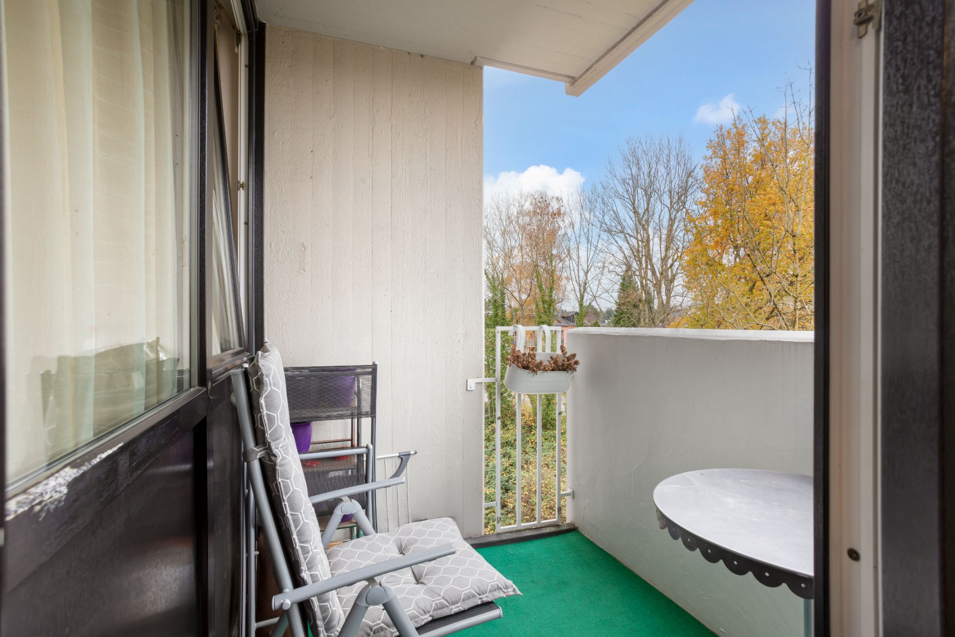 Kleiner Balkon mit zwei Stühlen, rundem Tisch und Blick auf Bäume im Herbst.