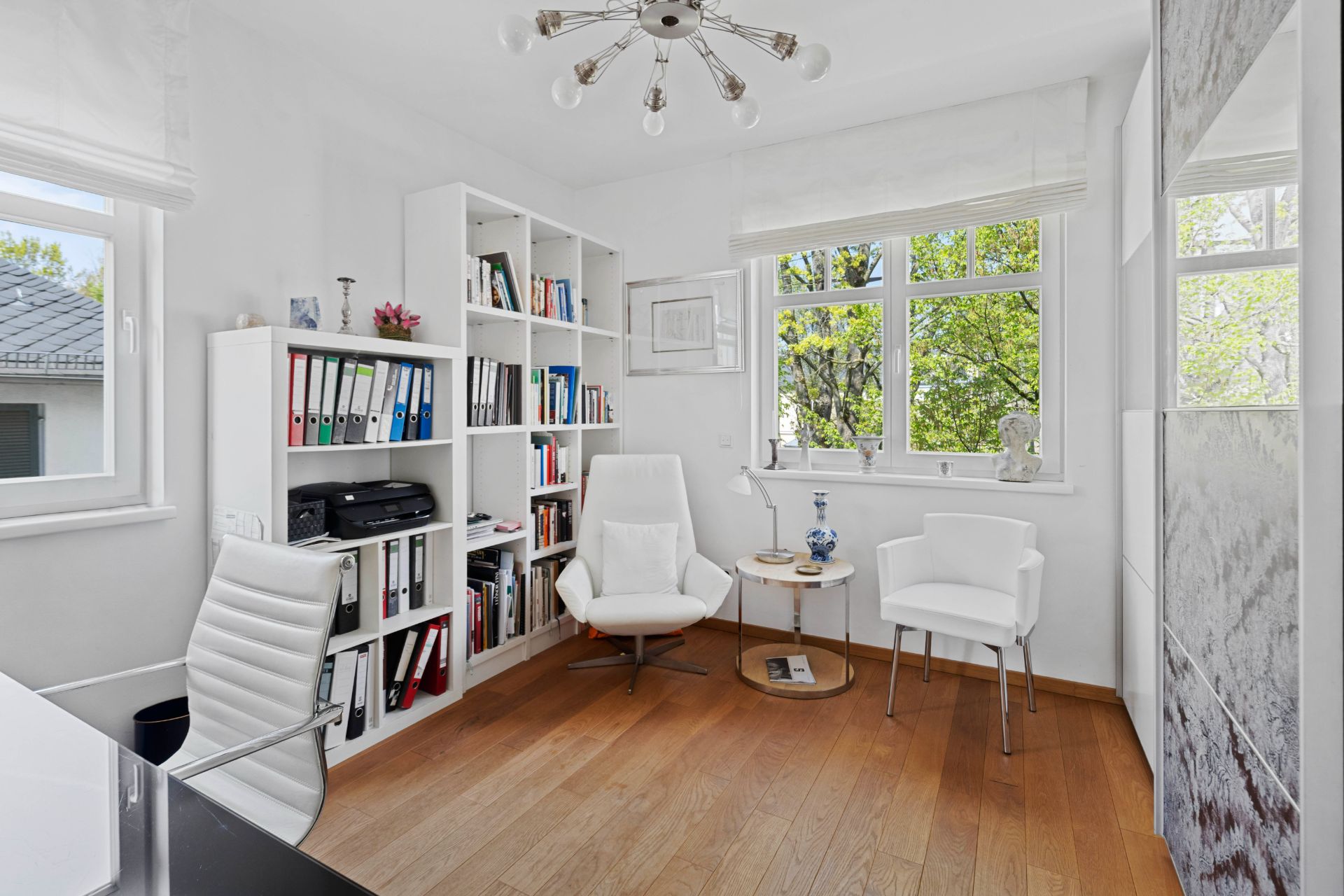 Helles Arbeitszimmer mit Holzfußboden, Bücherregal, Schreibtisch und zwei weißen Sesseln vor Fenstern.