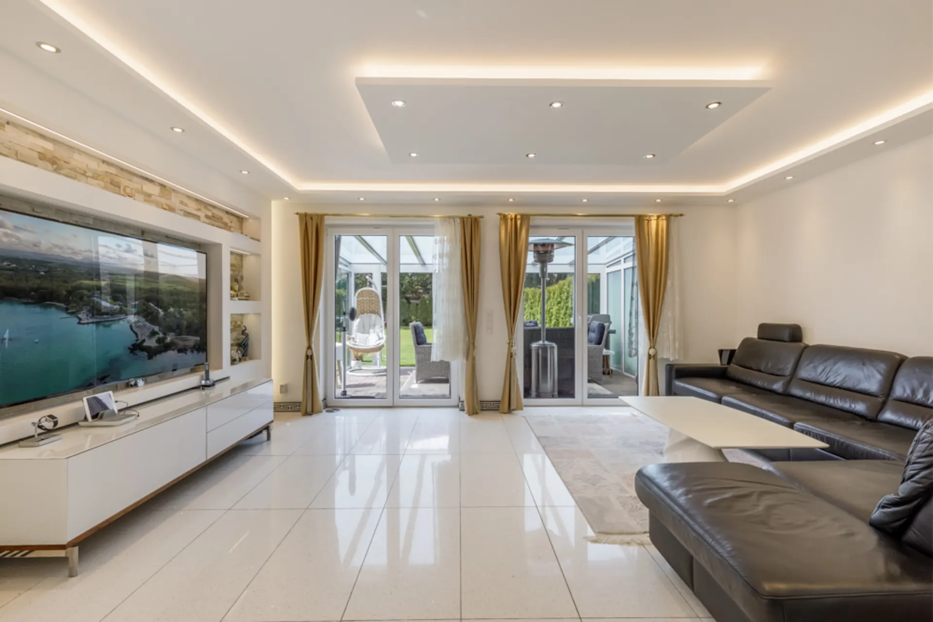 Modernes Wohnzimmer mit schwarzem Ledersofa, TV, großem Fenster und Zugang zur Terrasse.