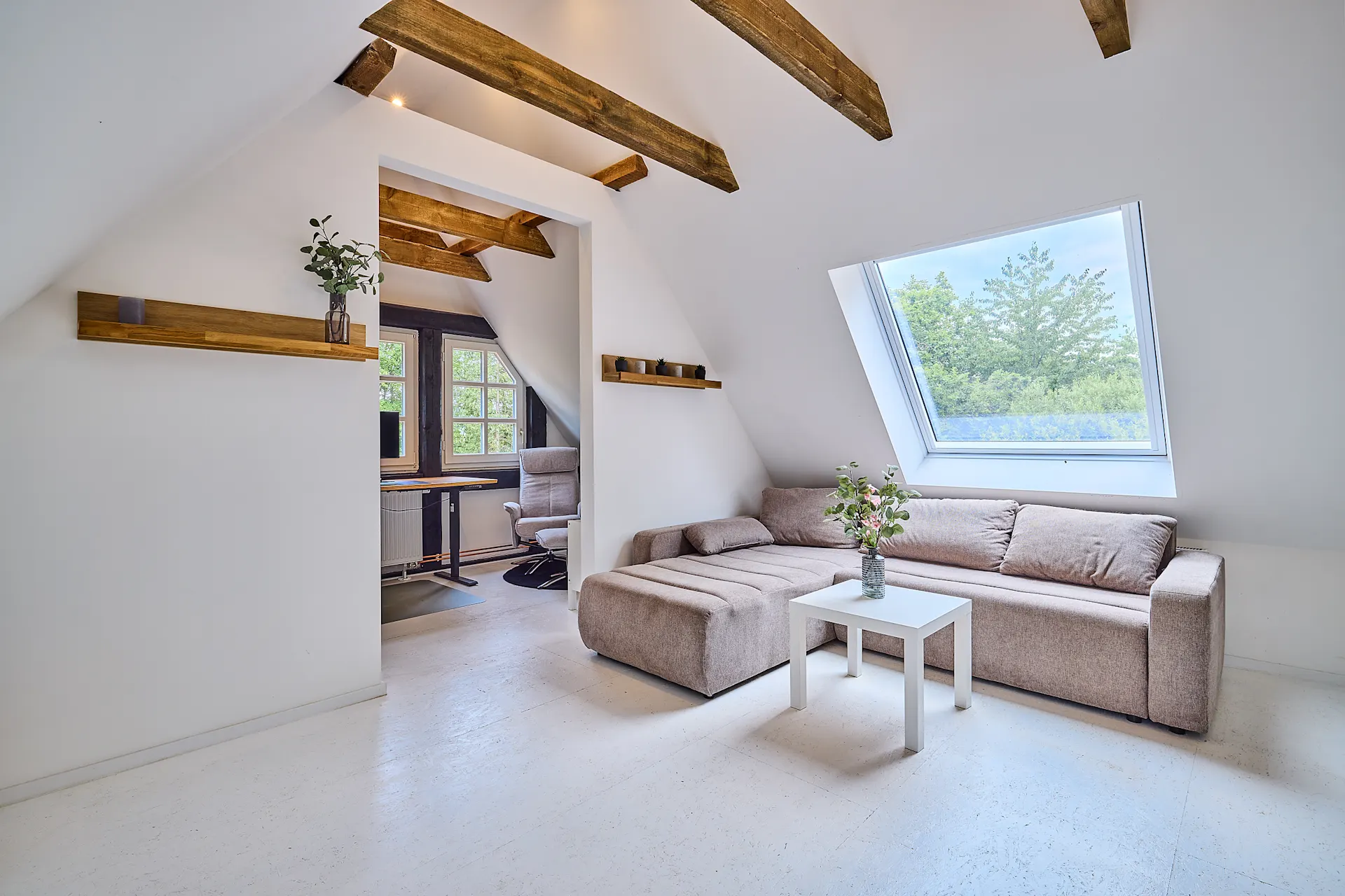 Helles Wohnzimmer mit Sofa, Couchtisch und Dachfenster, angrenzend an ein kleines Arbeitszimmer.