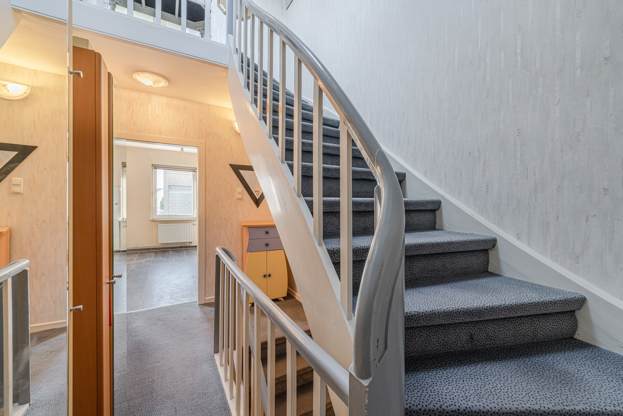Treppenhaus mit Teppichboden, Geländer und Zugang zu einem hellen Raum mit Fenster.