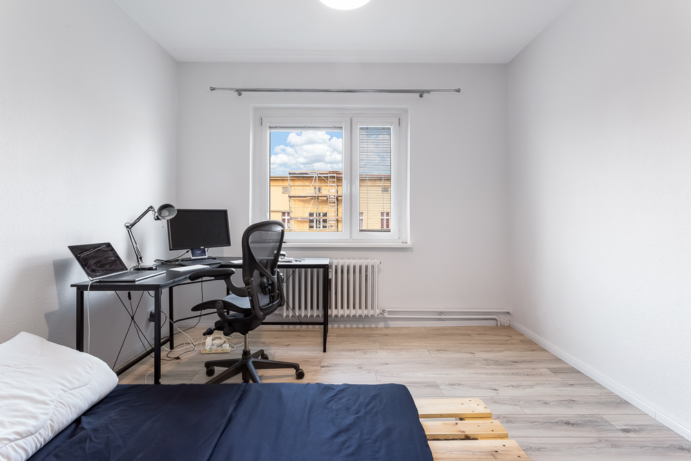 Helles Zimmer mit Schreibtisch, Bürostuhl, Bett und Fenster mit Blick nach draußen.