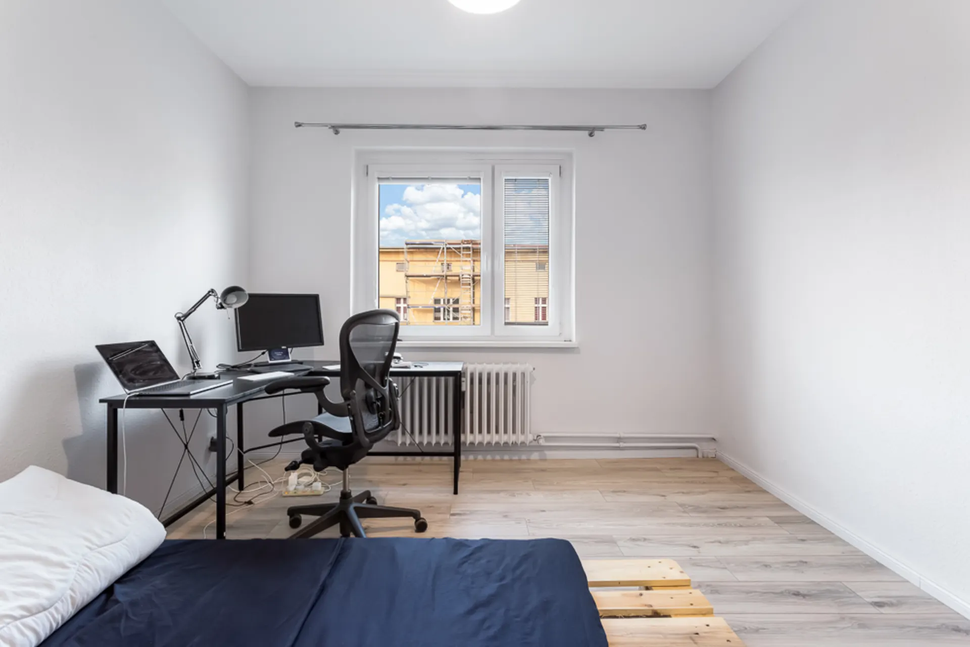 Helles Zimmer mit Schreibtisch, Bürostuhl, Bett und Fenster mit Blick nach draußen.