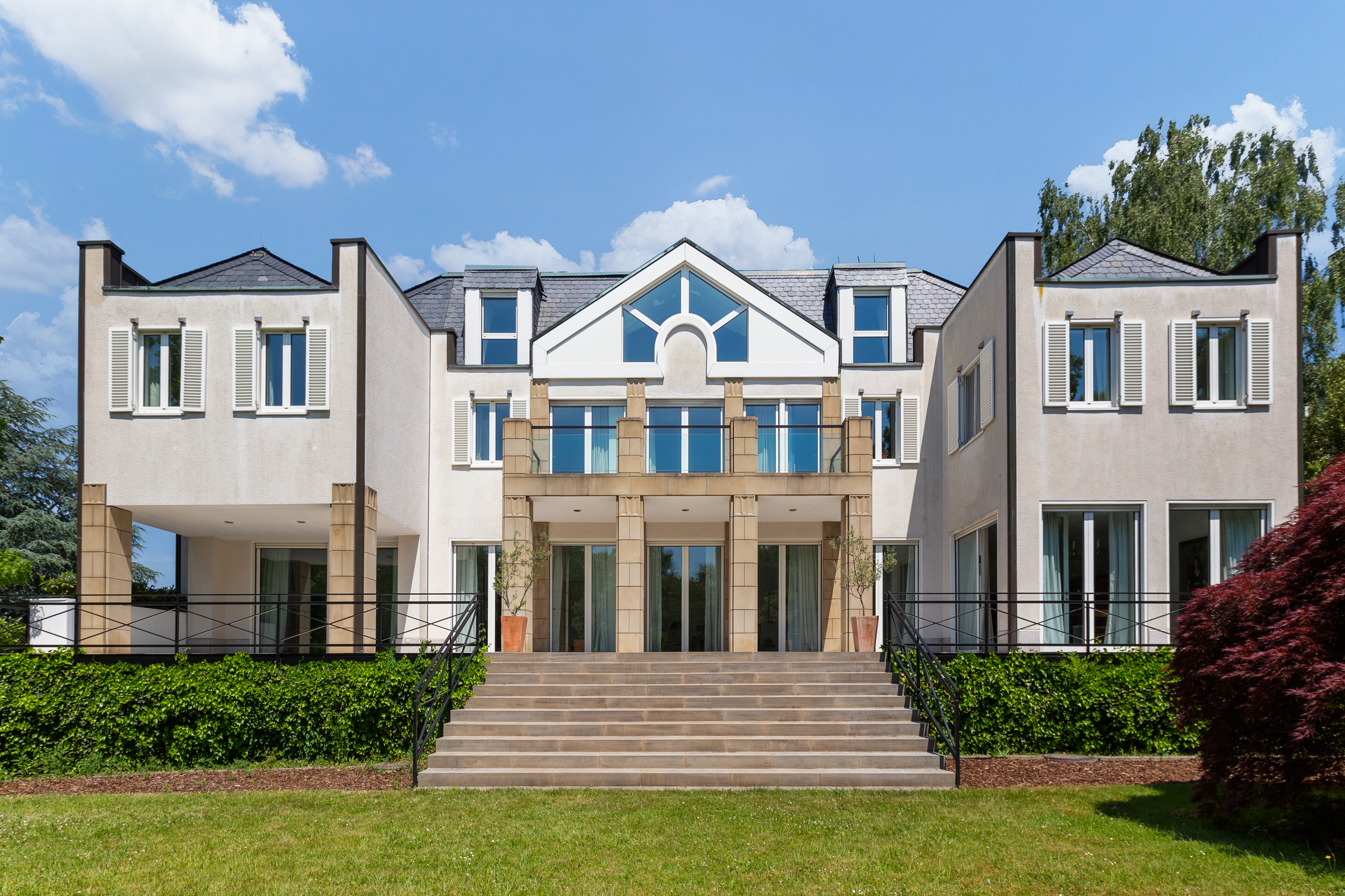 Großes, modernes Wohnhaus mit breiter Treppe, mehreren Fenstern und Balkon, umgeben von Garten.
