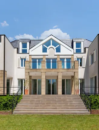 Großes, modernes Wohnhaus mit breiter Treppe, mehreren Fenstern und Balkon, umgeben von Garten.