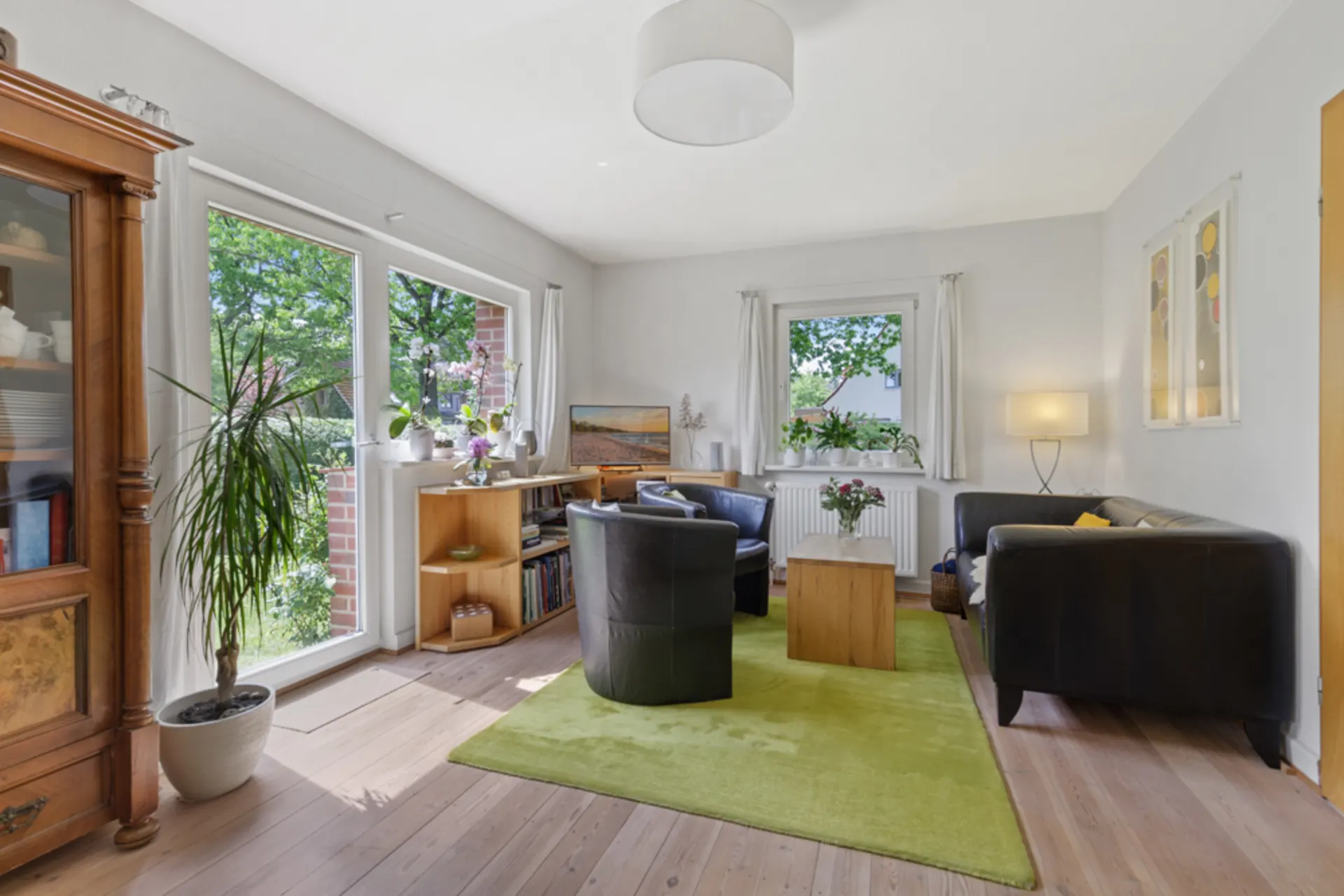 Helles Wohnzimmer mit großen Fenstern, zwei schwarzen Sofas, grünem Teppich und vielen Pflanzen.