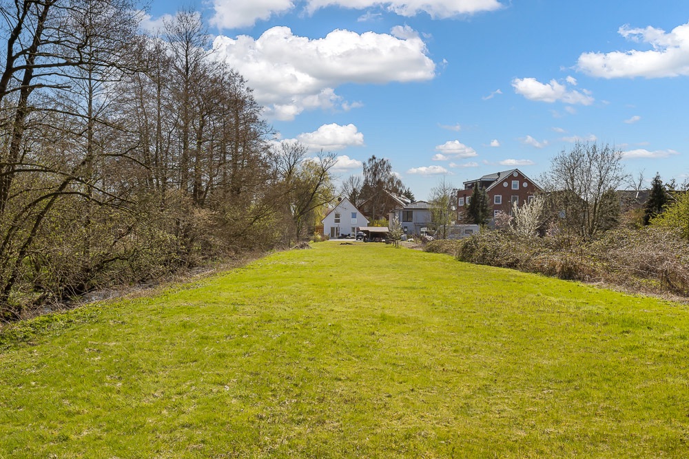 Große Rasenfläche mit Bäumen an den Seiten und Häusern im Hintergrund unter blauem Himmel.