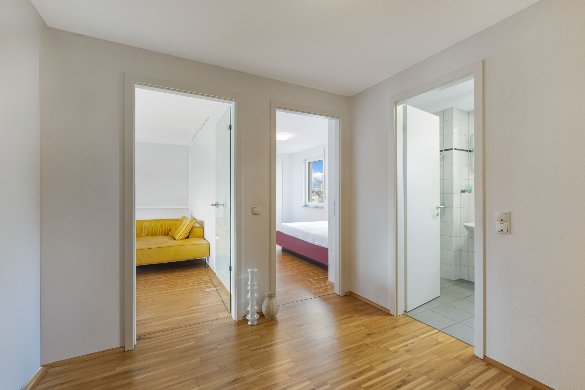 Flur mit drei Türen zu einem Wohnzimmer, Schlafzimmer und Badezimmer, Holzboden, weiße Wände.