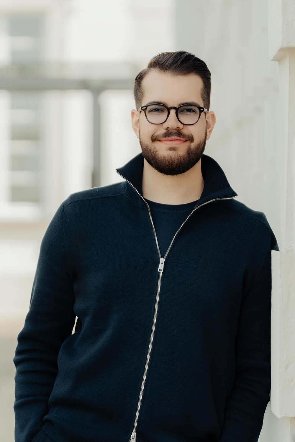 Mann mit Brille und Bart steht vor einem Gebäude mit Stein- und Fensterelementen.