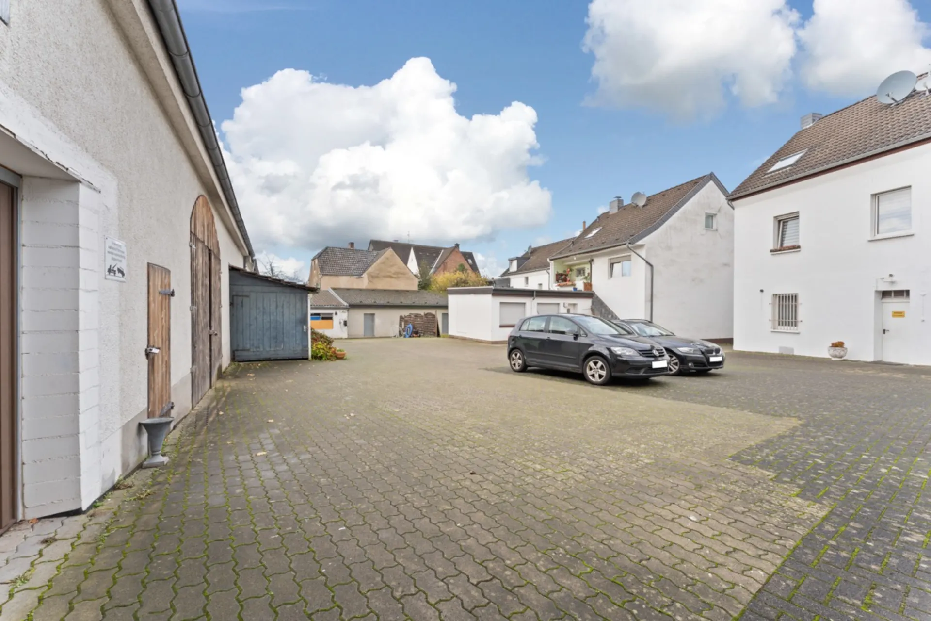 Gepflasterter Hof mit geparkten Autos, angrenzenden Garagen und Wohnhäusern im Hintergrund.