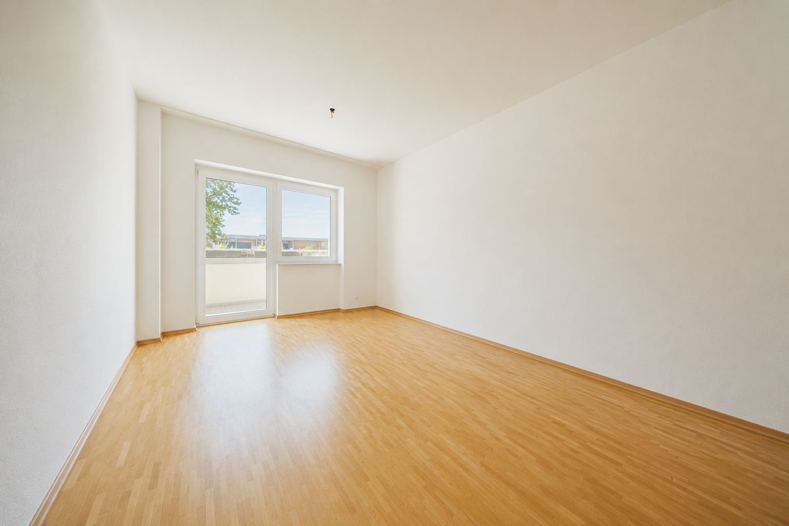Helles, leeres Zimmer mit Holzfußboden und großem Fenster mit Tür zum Balkon.