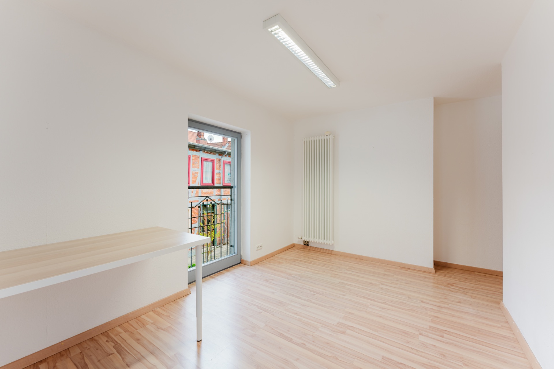 Helles Zimmer mit Holzboden, Tisch, Heizkörper und Tür zum Balkon.