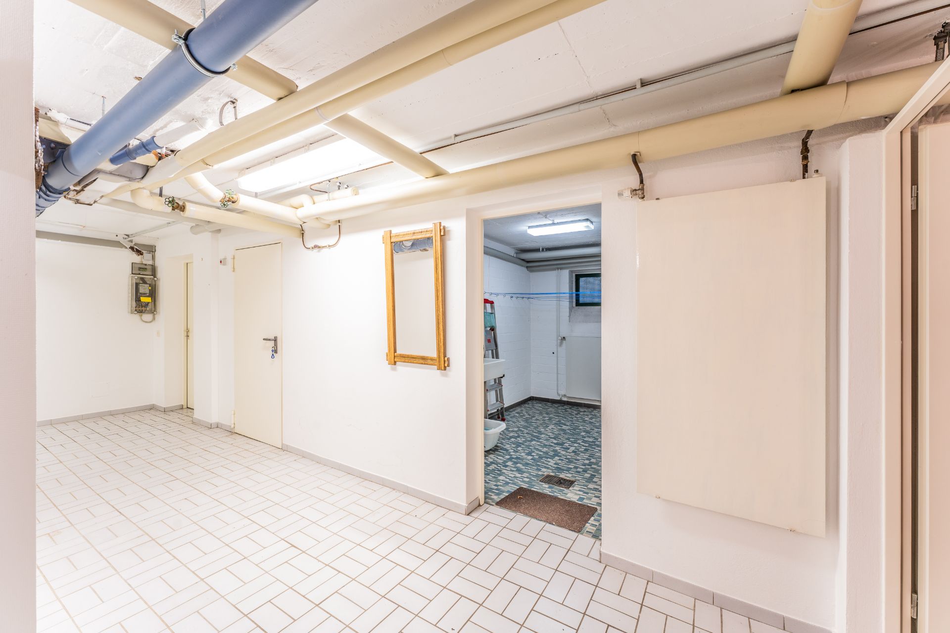 Heller Kellerraum mit weißen Fliesen, sichtbaren Rohren an der Decke und offenem Zugang zu einem zweiten Raum.