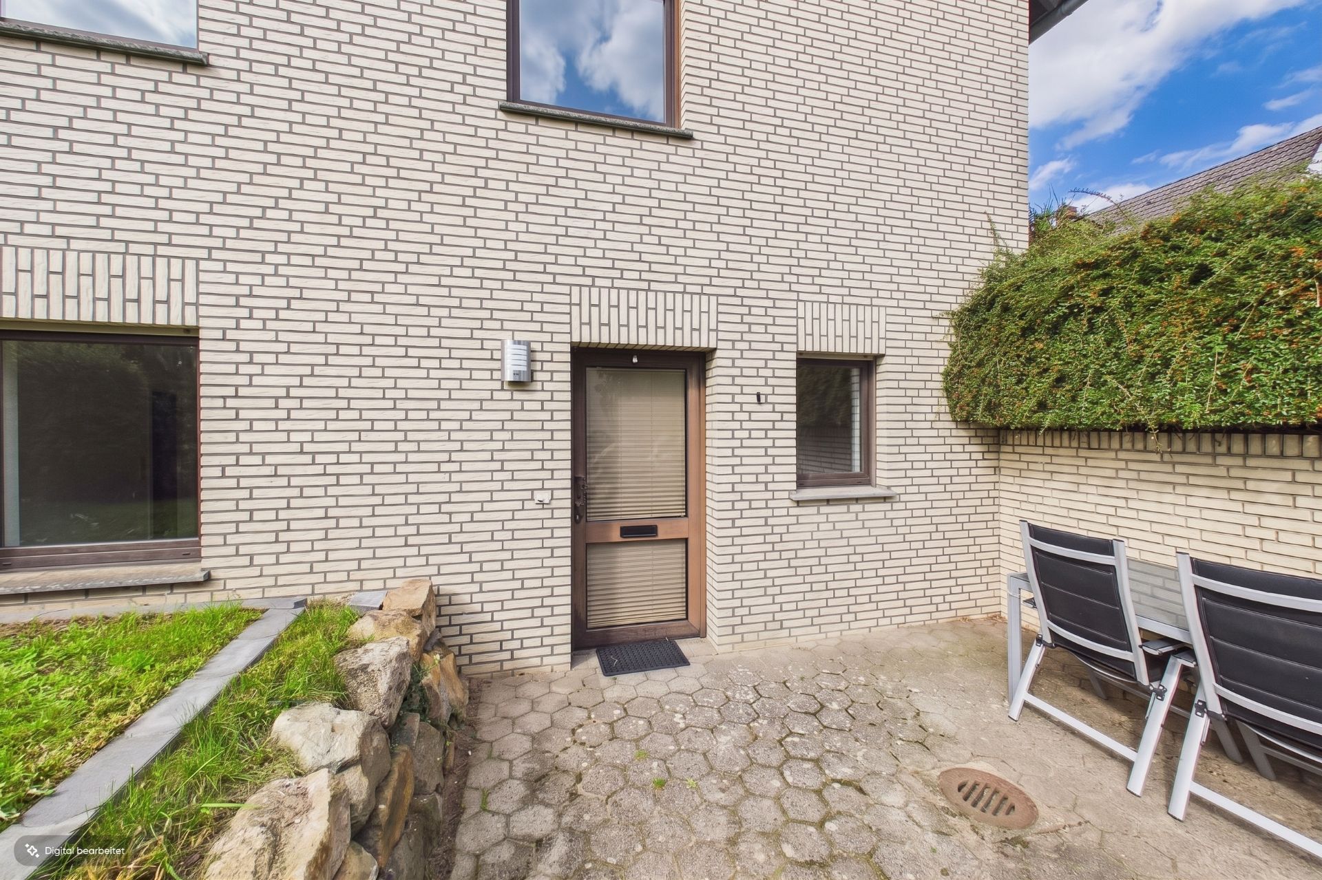 Hinterhof mit Pflastersteinen, moderner Haustür, Fenstern und mehreren Gartenstühlen an einer Mauer.