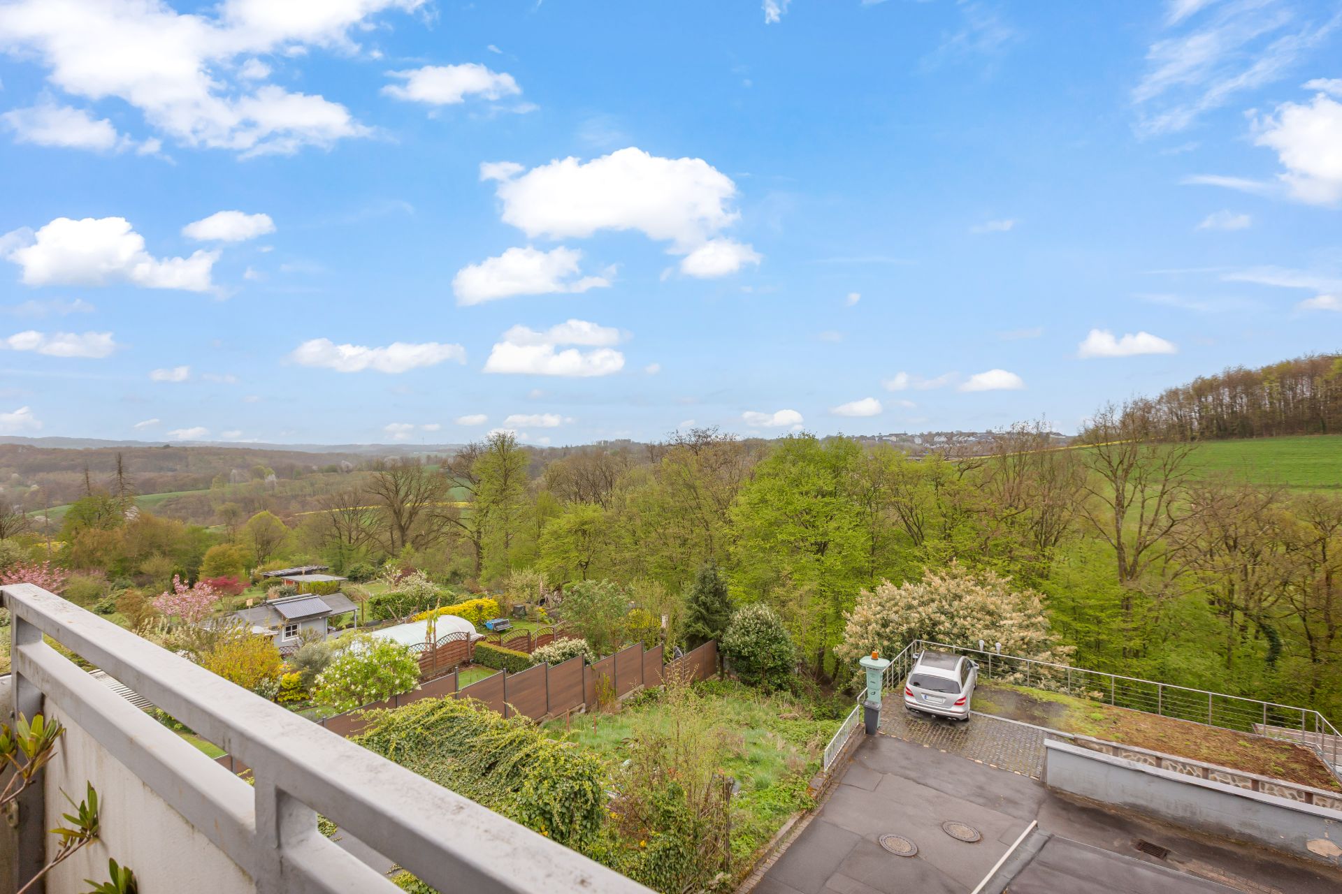 Blick von einem Balkon auf einen Garten, Bäume und ein geparktes Auto bei sonnigem Wetter.
