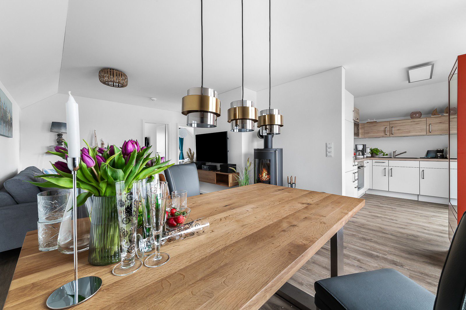 Ess- und Wohnzimmer mit Holztisch, Stühlen, Kamin, Küche im Hintergrund und Blumen auf dem Tisch.