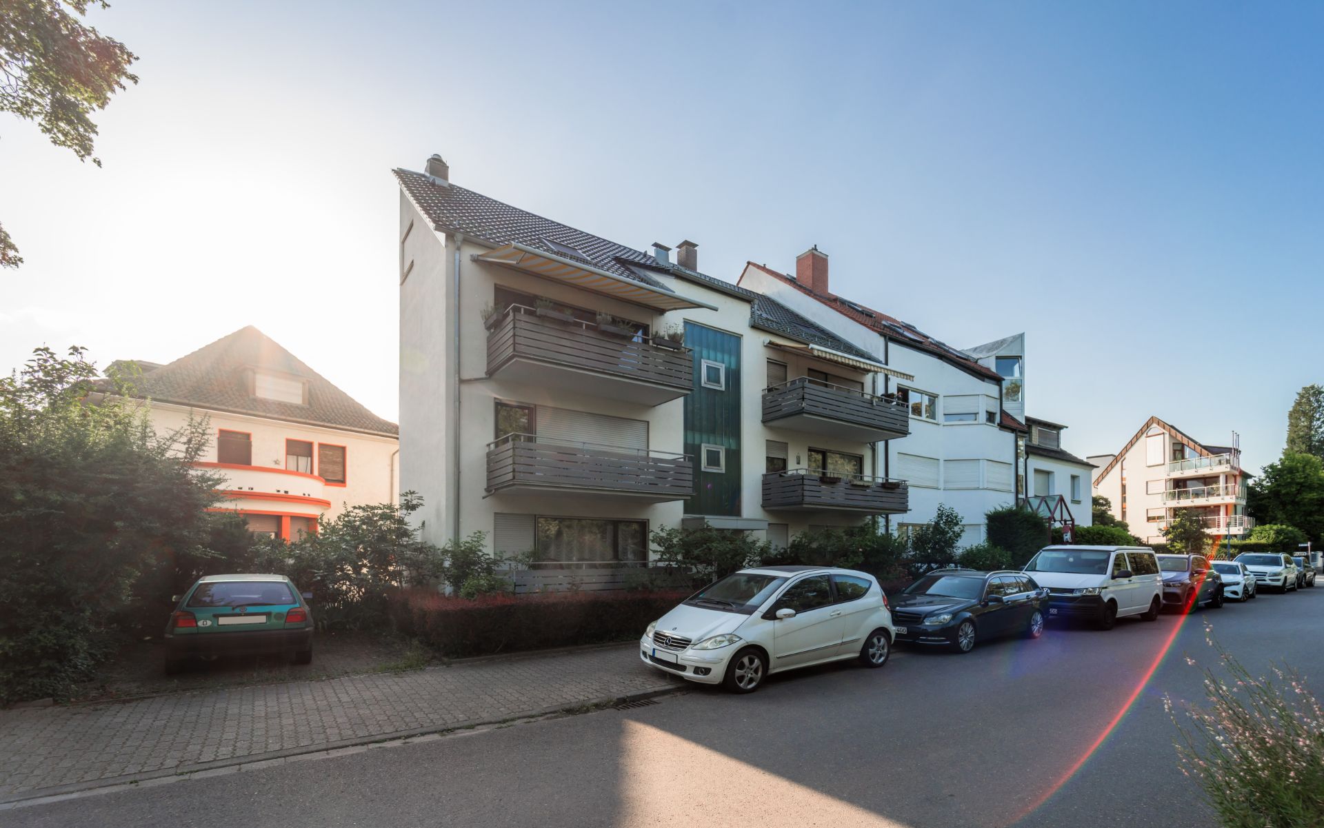 Mehrfamilienhaus mit Balkonen an einer ruhigen Straße, mehrere geparkte Autos davor.