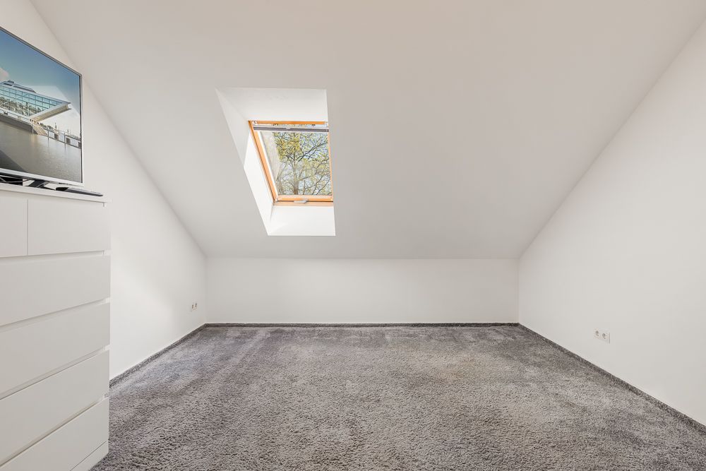 Helles Dachzimmer mit Teppichboden, Dachfenster und Kommode mit Fernseher.