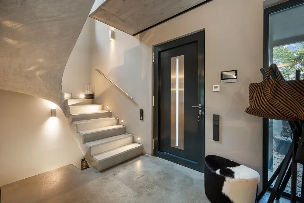 Moderner Eingangsbereich mit dunkler Tür, beleuchteter Treppe und Sitzgelegenheit.