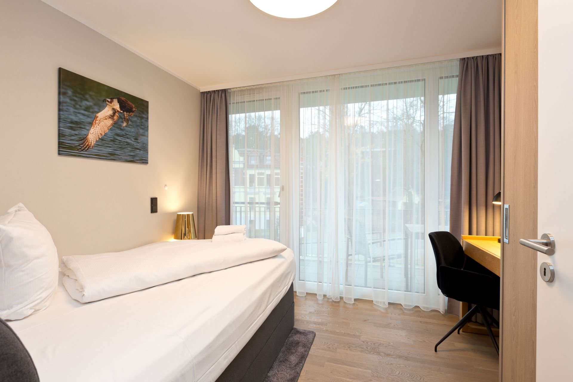 Helles Schlafzimmer mit Einzelbett, Schreibtisch und großen Fenstern mit Vorhängen.