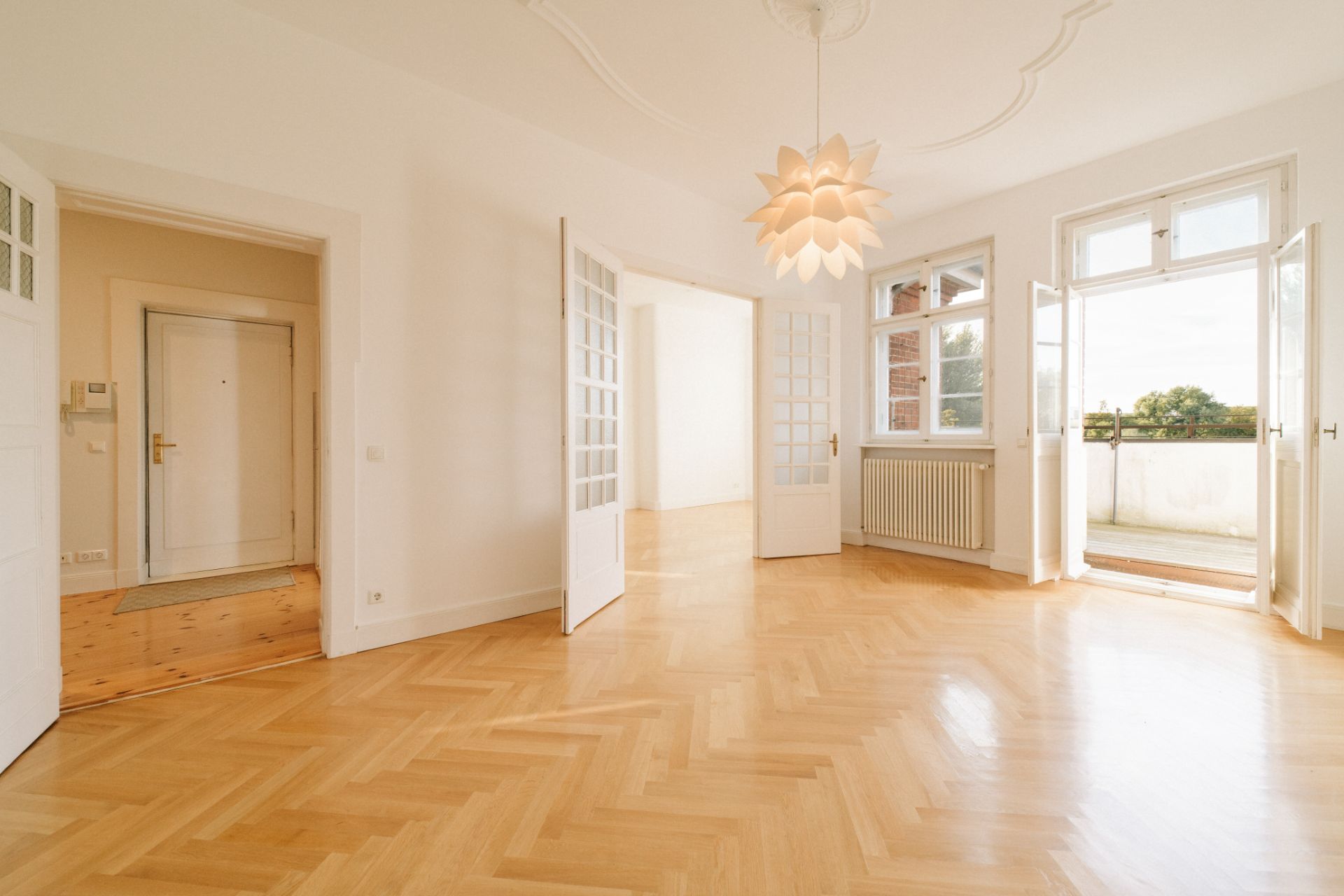 Helles Wohnzimmer mit Holzparkett, offenen Türen, Fenster, Heizung und Zugang zum Balkon.