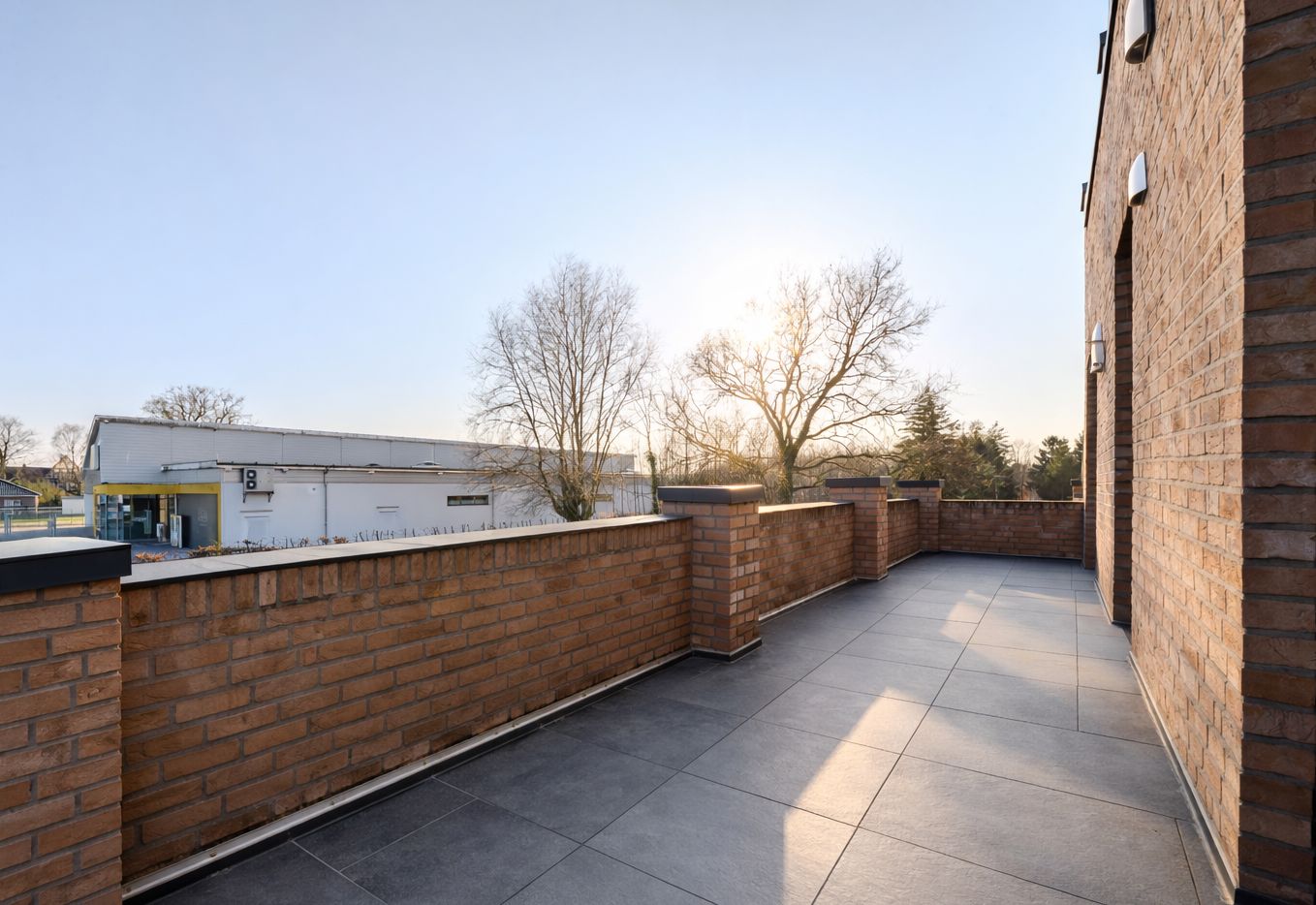 Große Terrasse mit Fliesenboden und gemauerter Brüstung, daneben Bäume und Gebäude im Hintergrund.