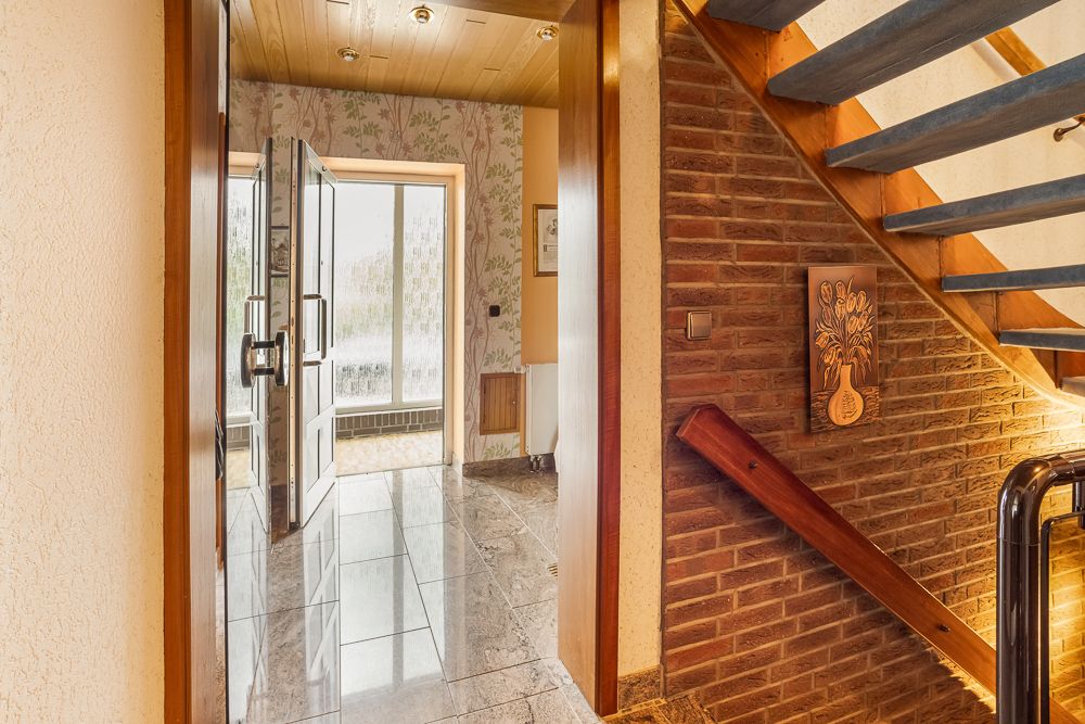 Flur mit Treppe, Fliesenboden und Glastür, gemusterten Wänden und Holzdecke.