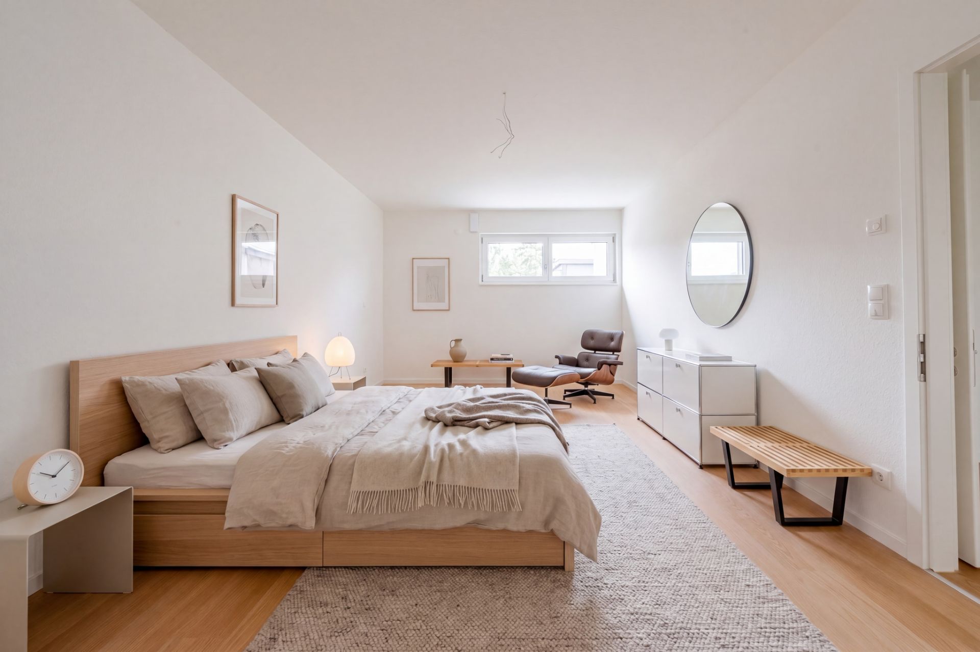 Helles Schlafzimmer mit Doppelbett, Sessel, Kommode, rundem Spiegel und großem Teppich.