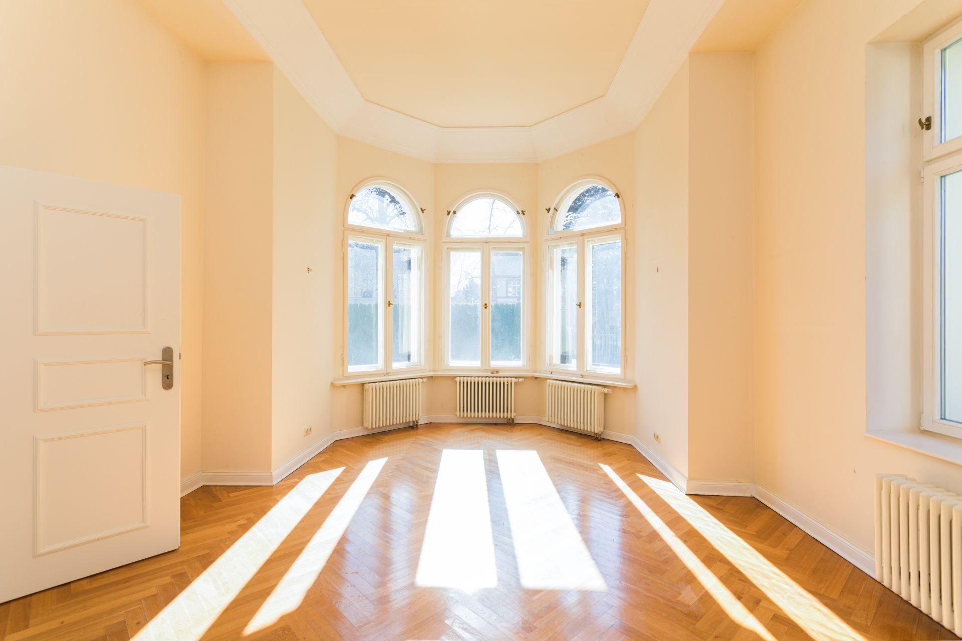 Helles Zimmer mit Parkettboden, mehreren Fenstern und weißer Tür, Sonnenlicht fällt hinein.