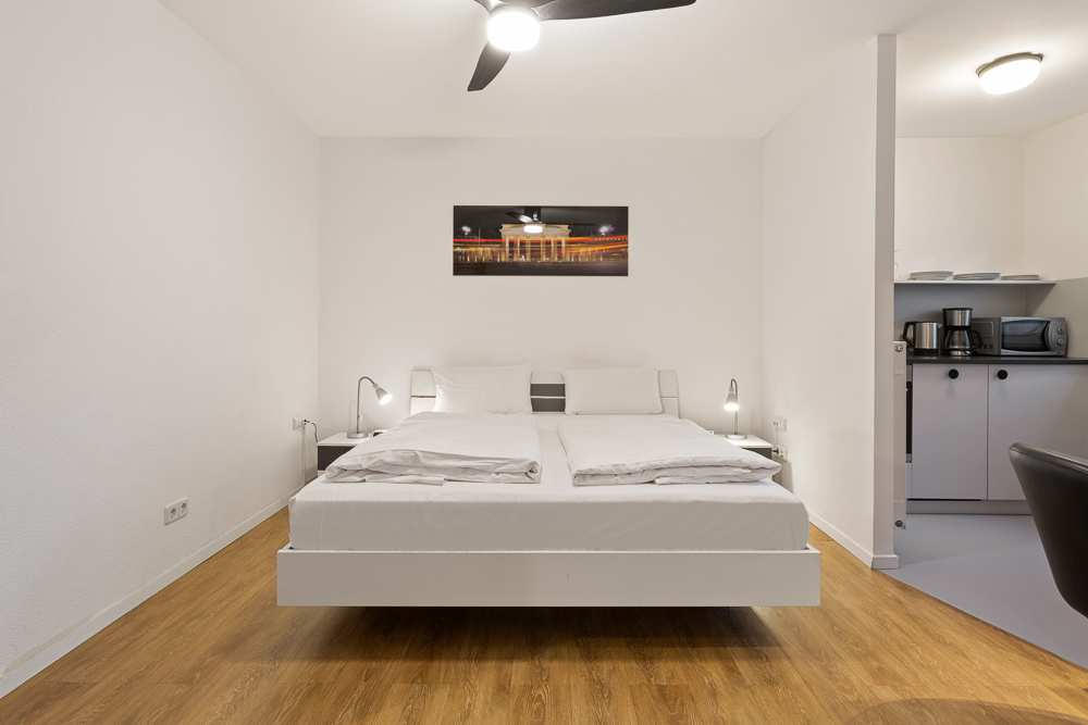 Schlafzimmer mit Doppelbett, zwei Nachttischen, Lampen und Blick auf eine kleine Küchenzeile.