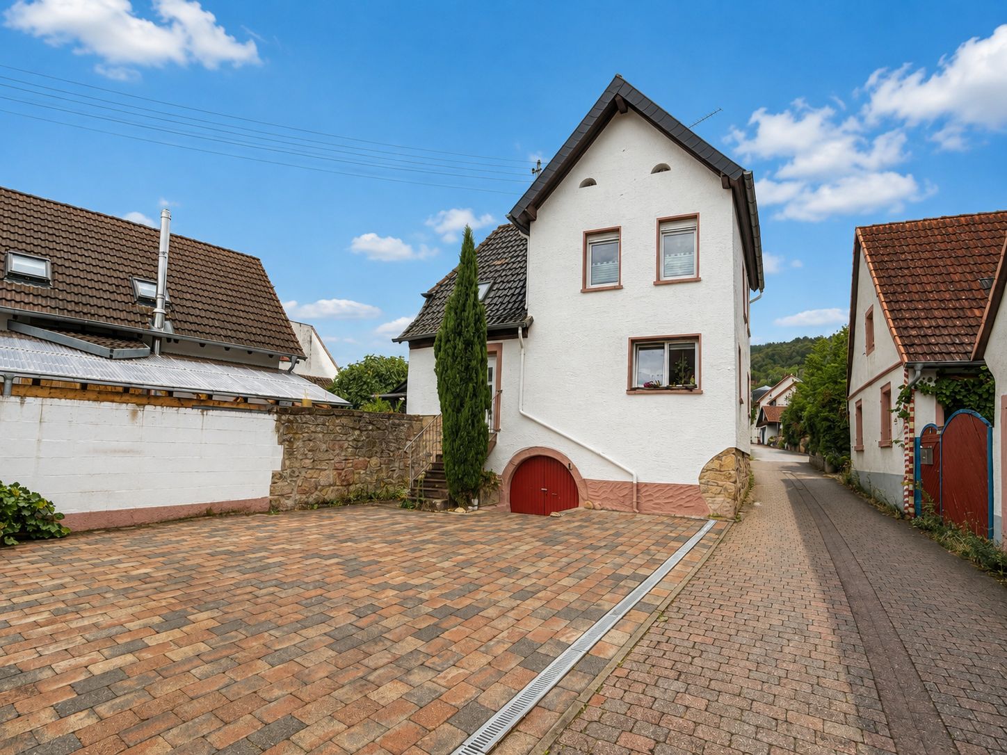 Weißes Einfamilienhaus mit rotem Garagentor und gepflastertem Hof, daneben eine schmale Straße.
