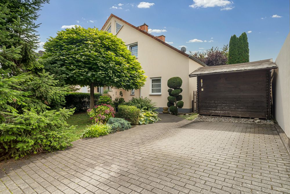 Gepflegter Garten mit Bäumen, Sträuchern und gepflasterter Einfahrt vor einem Wohnhaus.