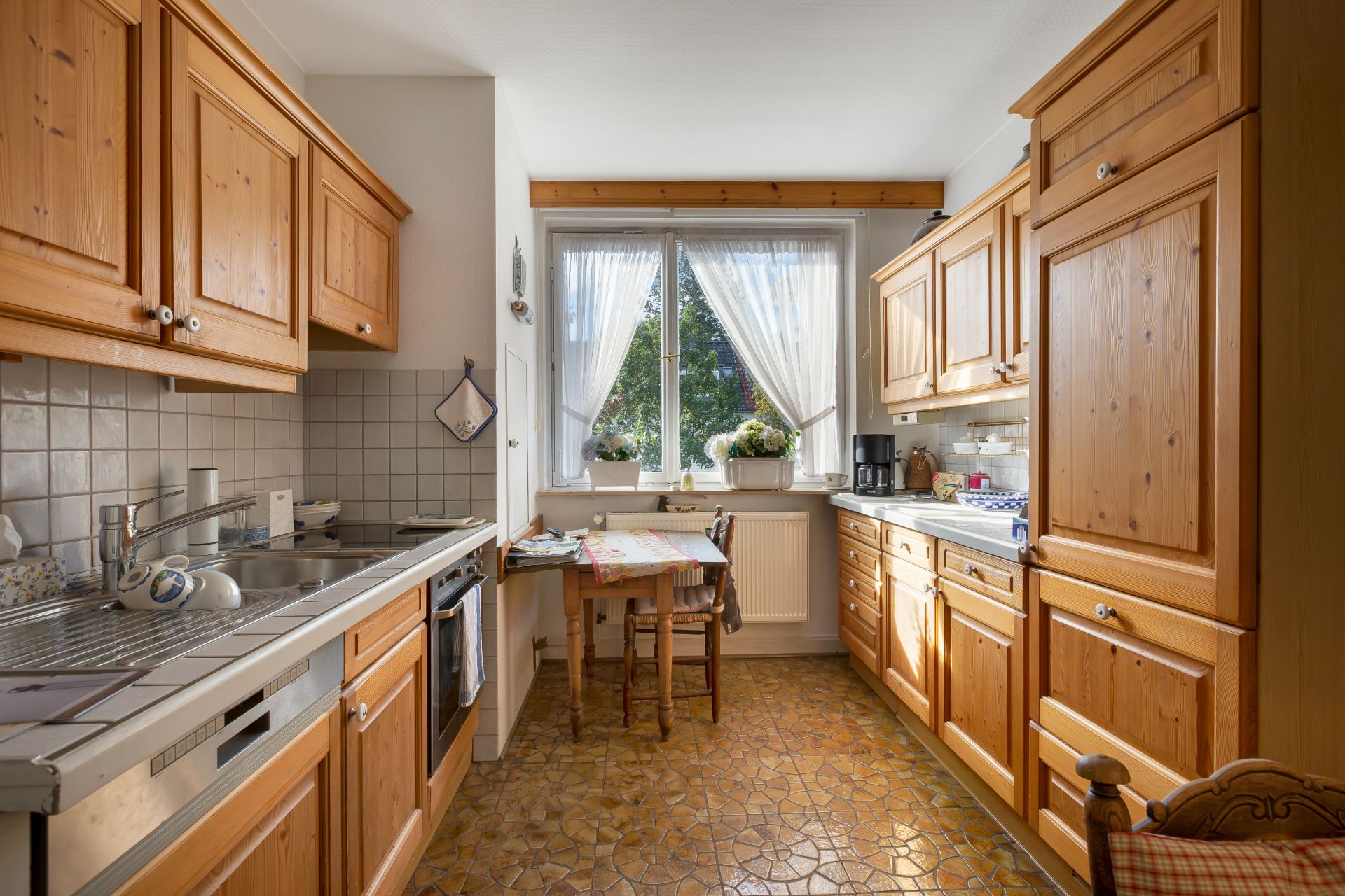 Helle Küche mit Holzmöbeln, kleinem Esstisch und großem Fenster mit weißen Gardinen.
