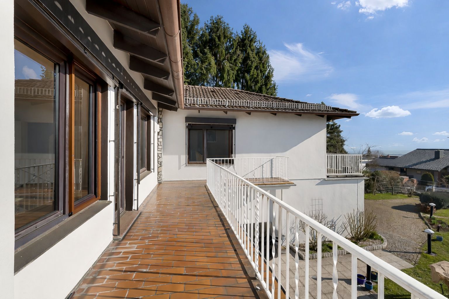 Langer Balkon mit braunen Fliesen und weißem Geländer an einem Wohnhaus, Ausblick auf Garten und Nachbarhäuser.