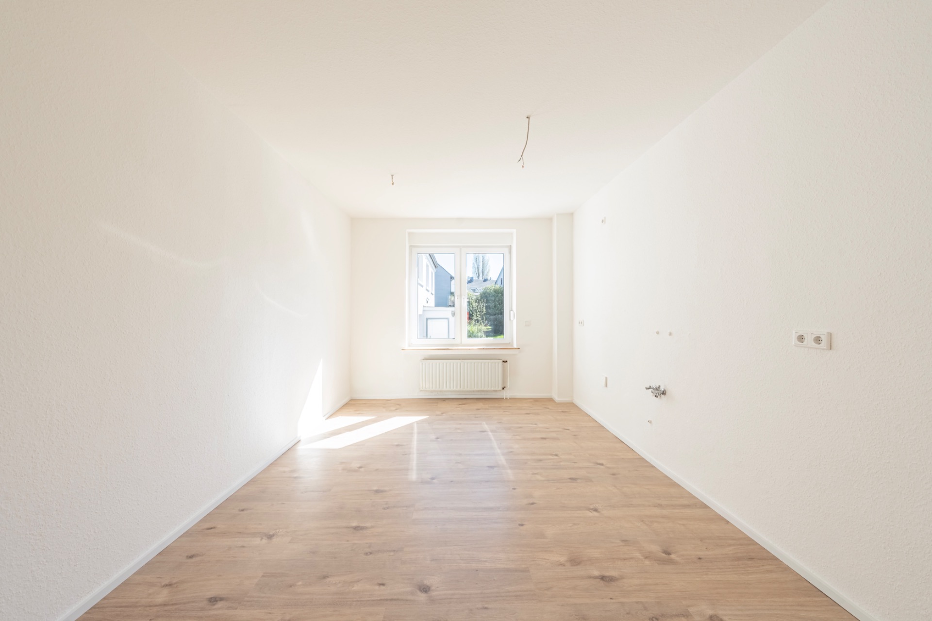 Heller, leerer Raum mit Holzfußboden und Fenster am Ende, darunter ein Heizkörper.