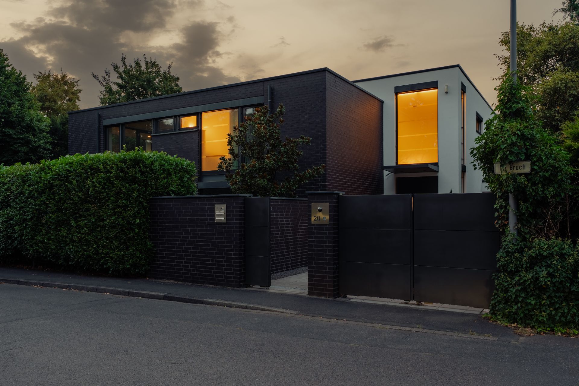 Moderne zweistöckige Wohnhausfassade mit großen Fenstern und Gartenhecke bei Abenddämmerung.