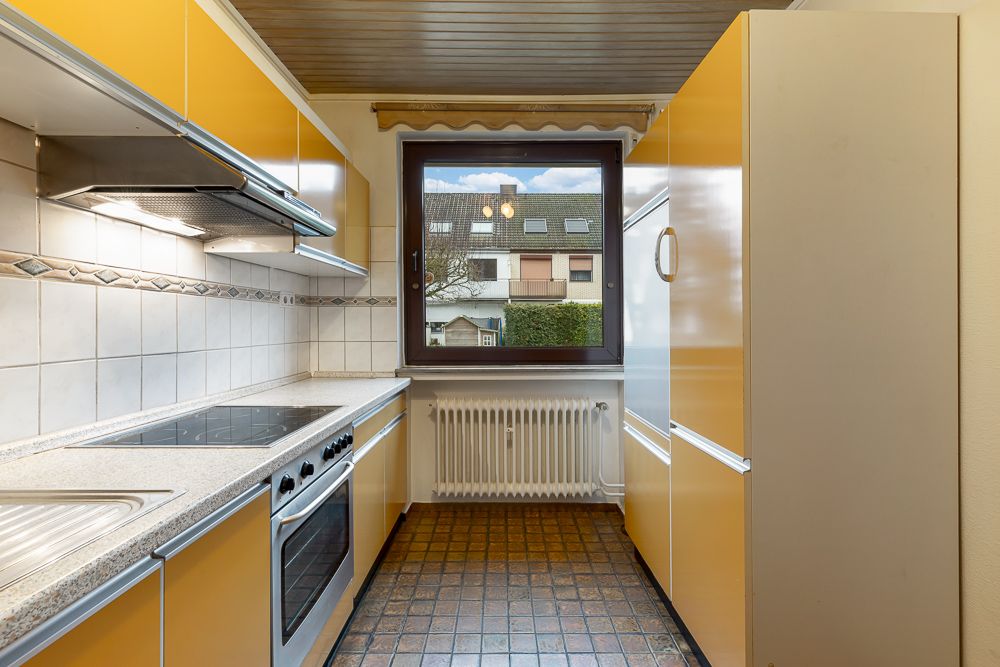 Schmale Küche mit gelben Schränken, Elektroherd, Fenster und Blick auf ein Wohnhaus.