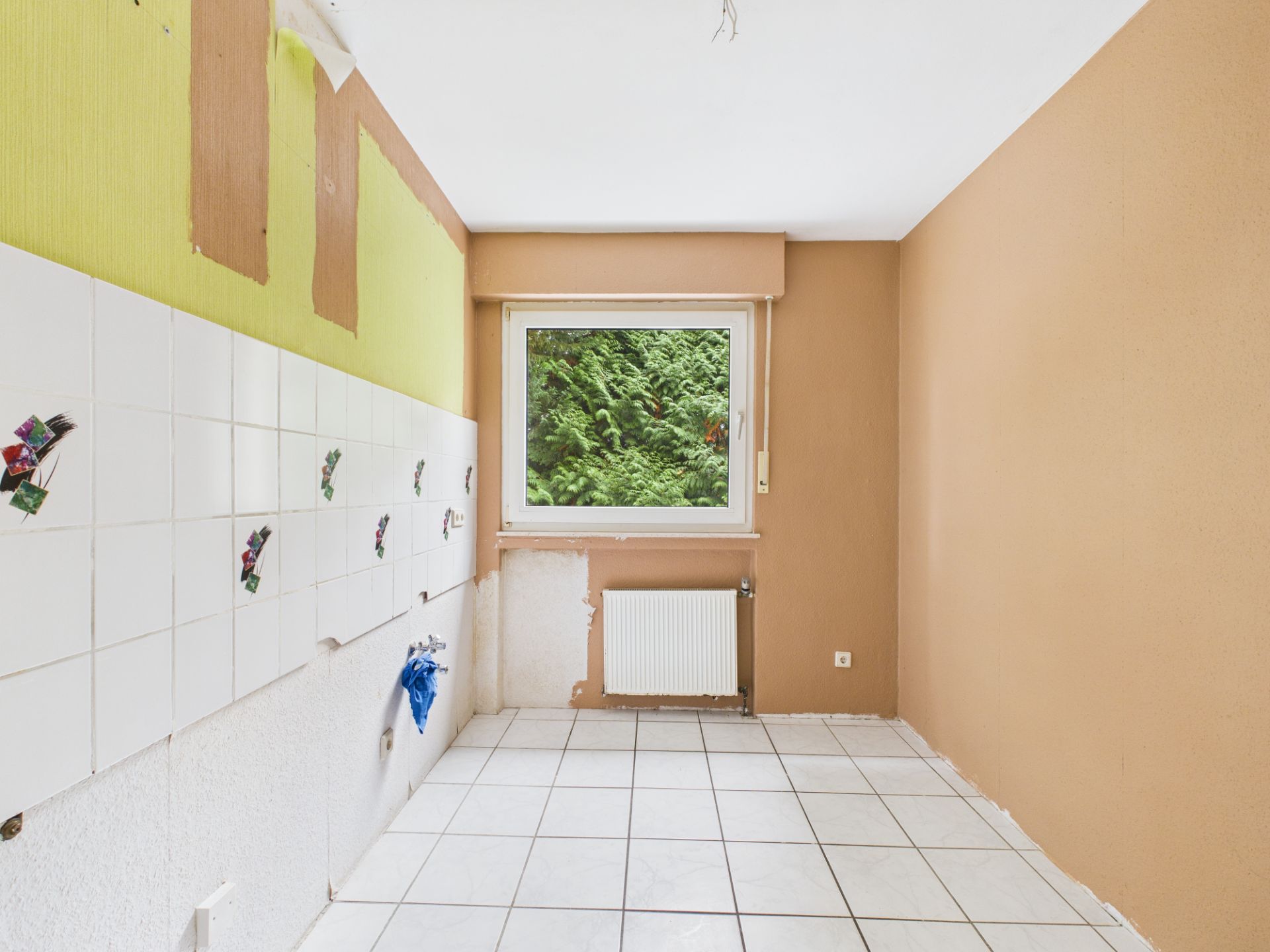 Leerer Raum mit gefliestem Boden, braunen Wänden und einem Fenster mit Blick auf Grün.