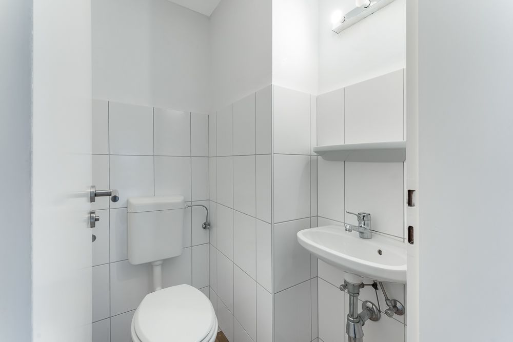 Kleines Badezimmer mit WC, weiß gefliest, kleinem Waschbecken und Spiegel mit Licht.