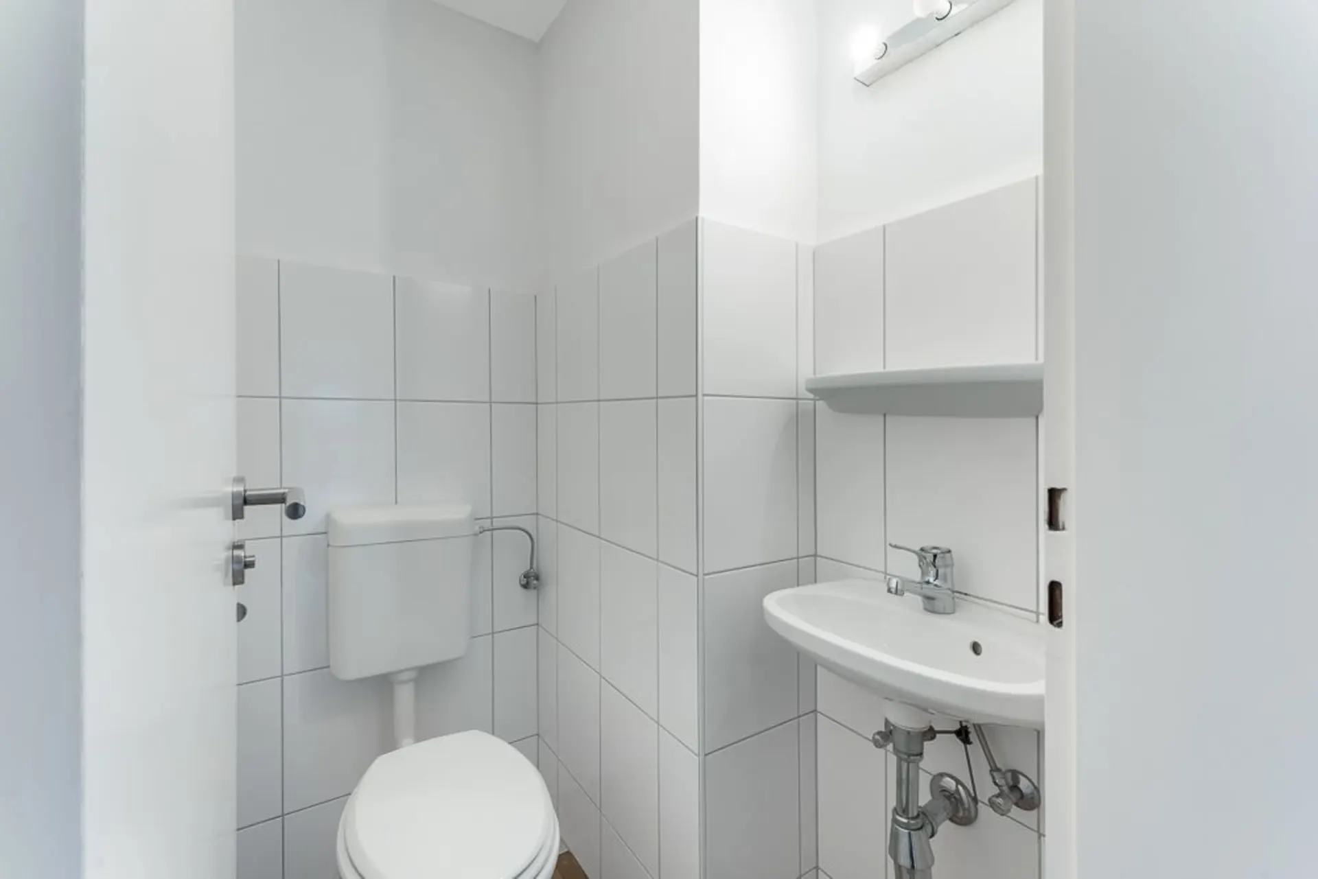 Kleines Badezimmer mit WC, weiß gefliest, kleinem Waschbecken und Spiegel mit Licht.