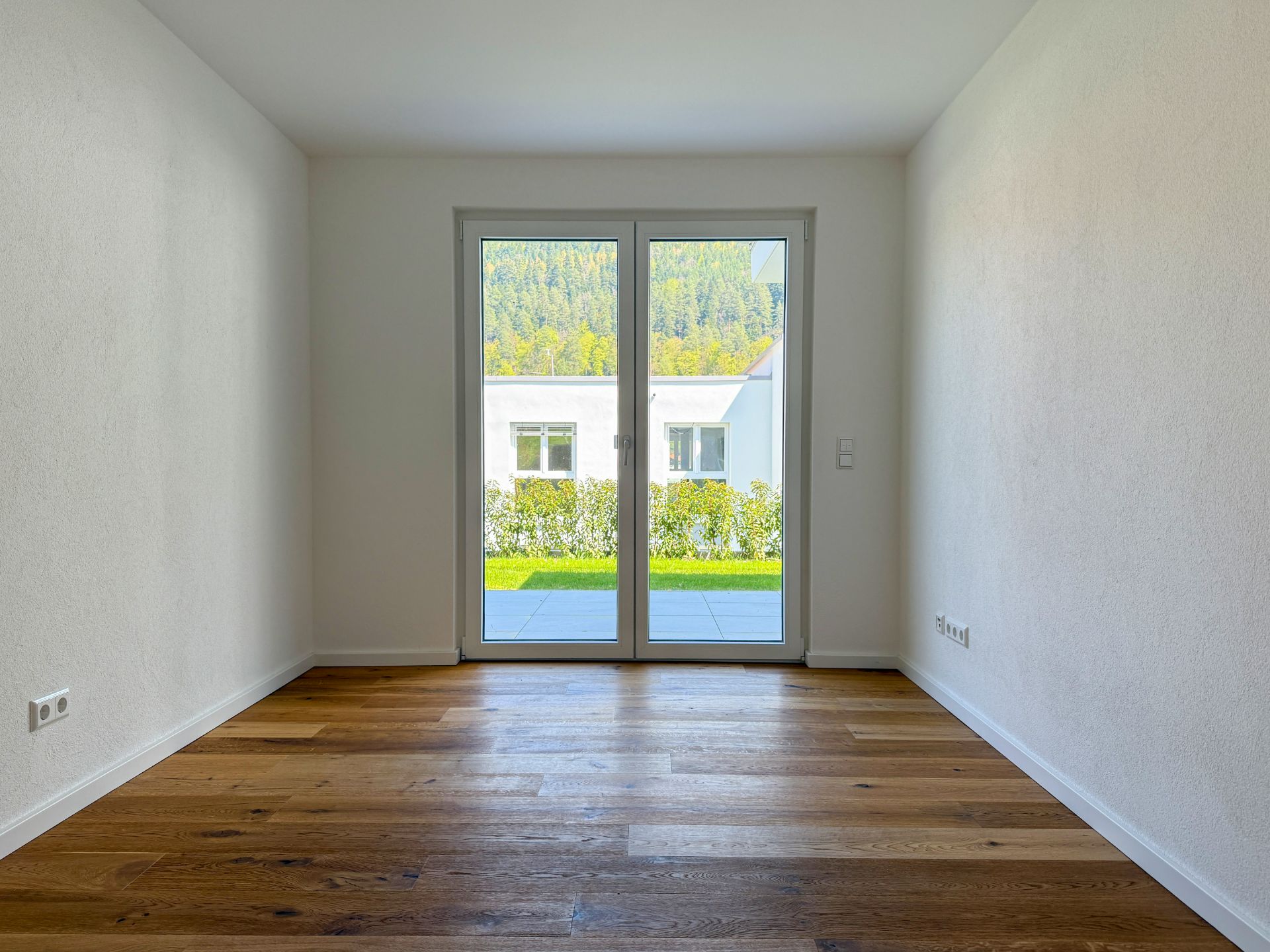 Leerer Raum mit Holzfußboden und großen Glastüren, die auf eine Terrasse und Garten führen.