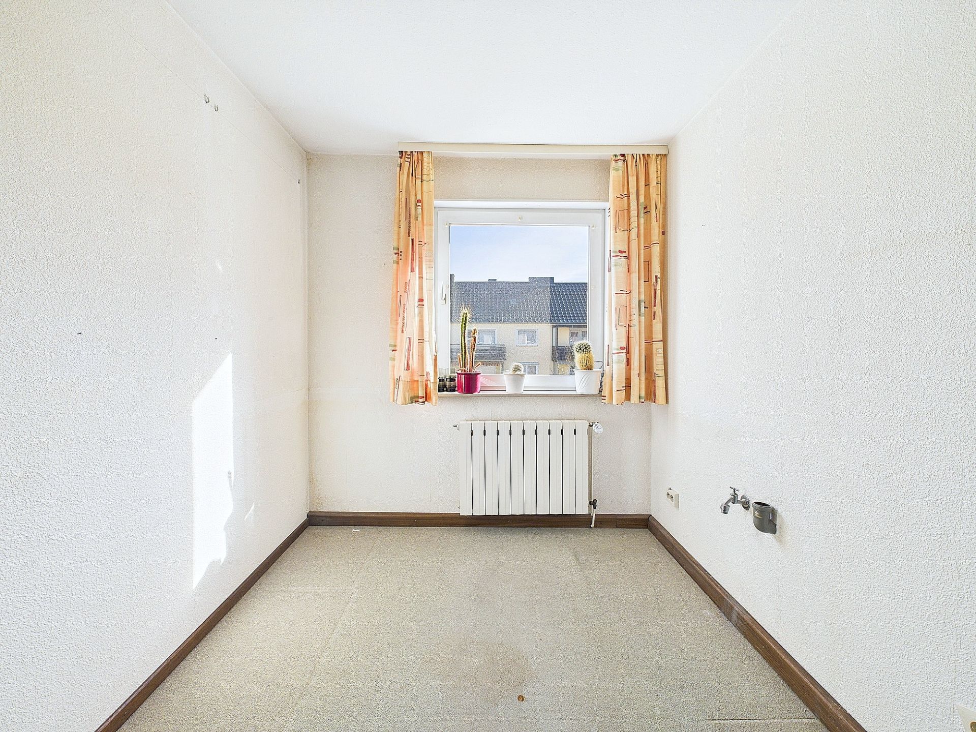 Leeres Zimmer mit Fenster, gelben Vorhängen, Heizkörper und zwei kleinen Pflanzen auf der Fensterbank.