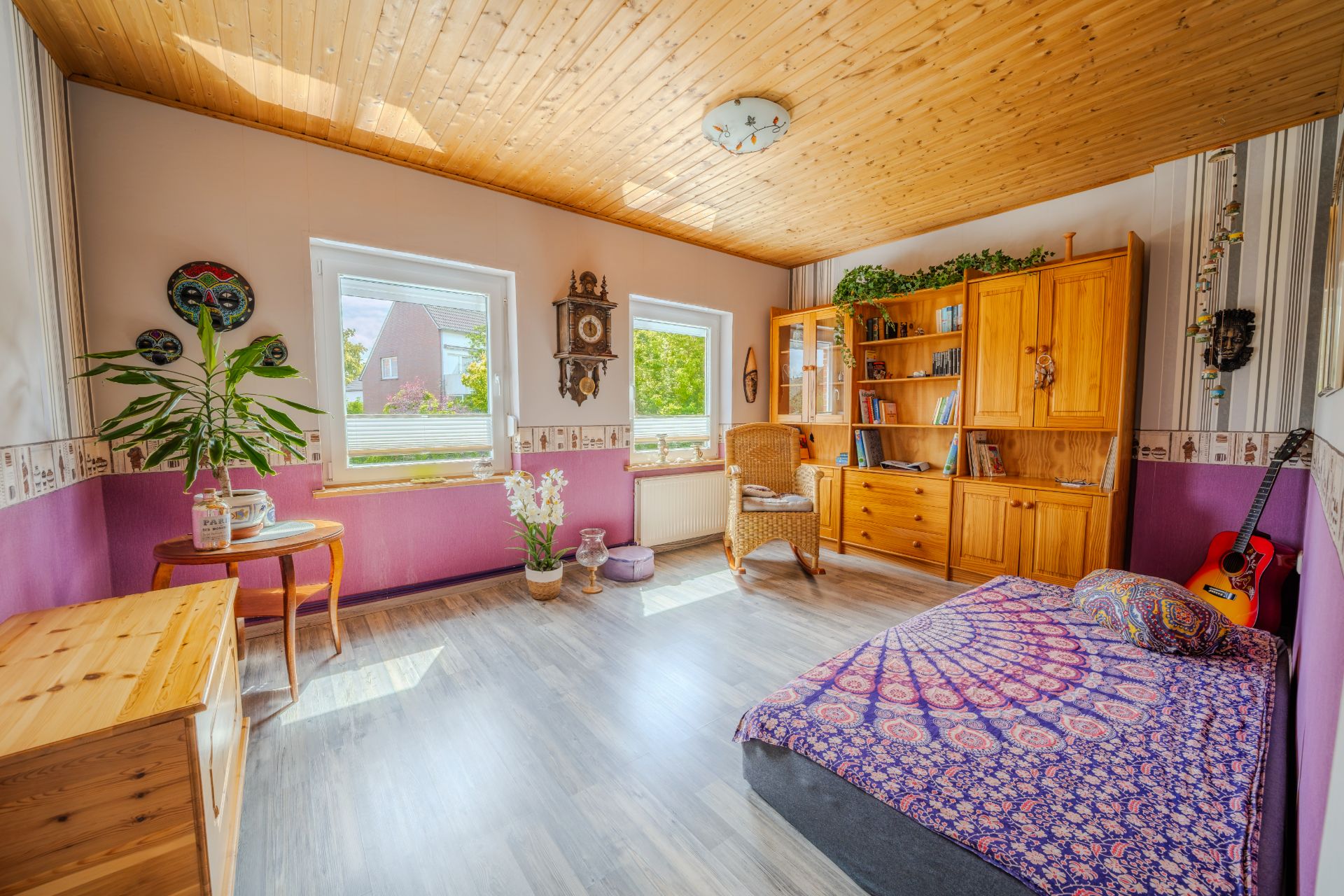 Helles Zimmer mit Holzmöbeln, Bett, Pflanzen, einer Gitarre und großen Fenstern.