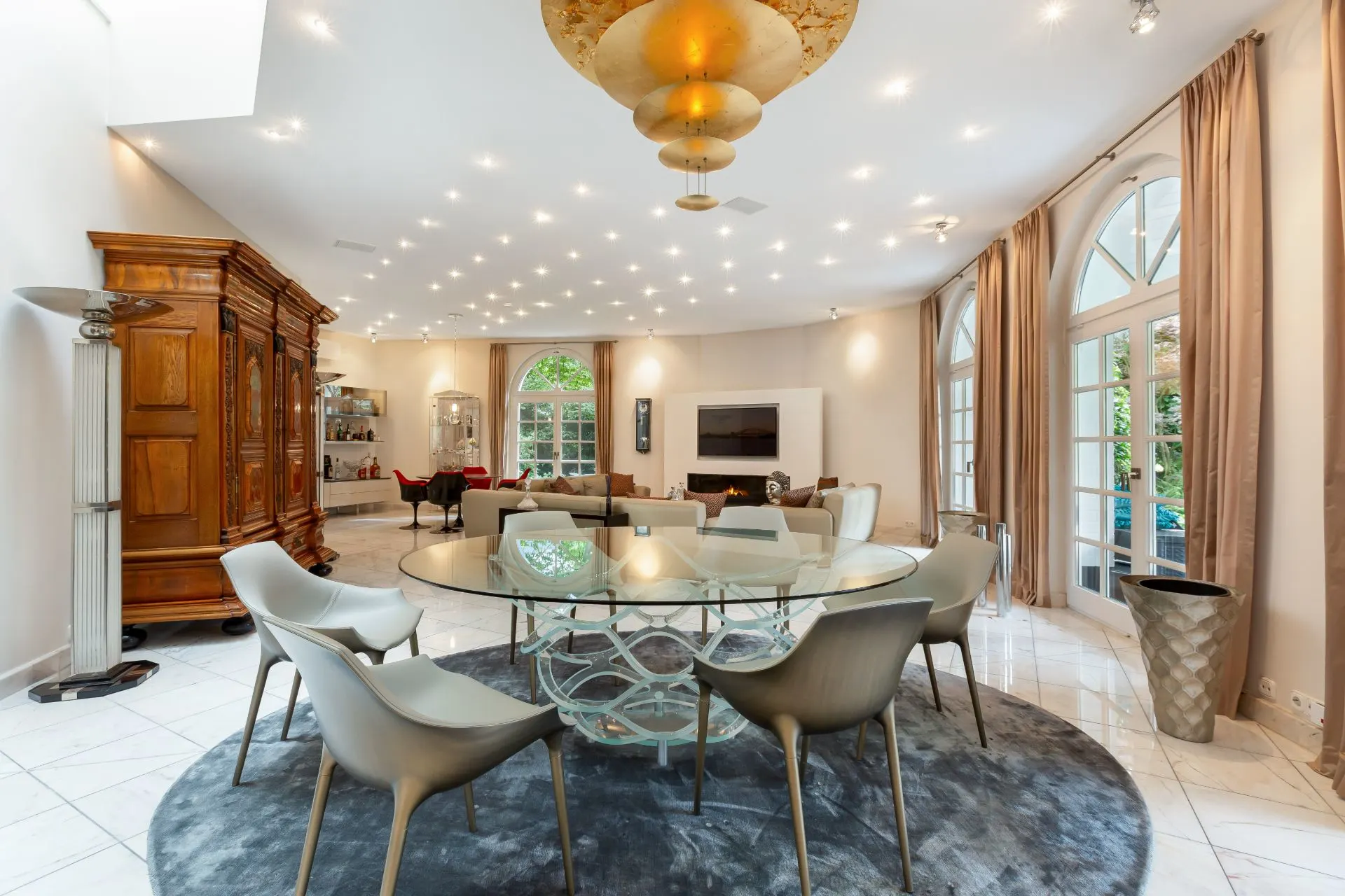 Modernes Wohnzimmer mit rundem Esstisch, sechs Stühlen, großen Fenstern und Holzmöbeln.