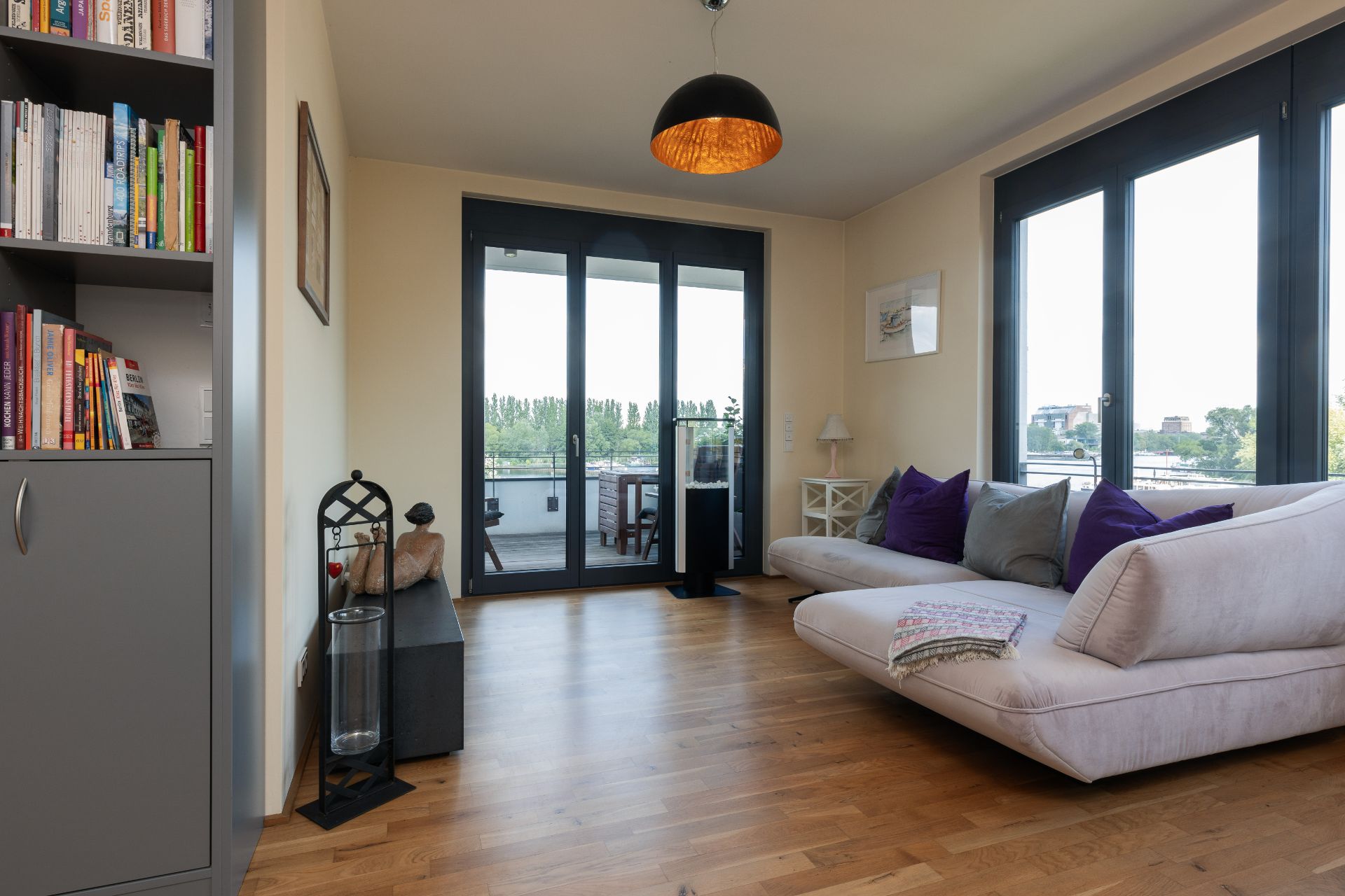 Wohnzimmer mit großem Sofa, Bücherregal, Holzfußboden und Fenstern zu einem Balkon.