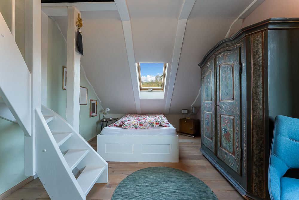 Schlafzimmer mit Bett unter Dachschräge, Fenster, großem Schrank und blauer Sitzgelegenheit.