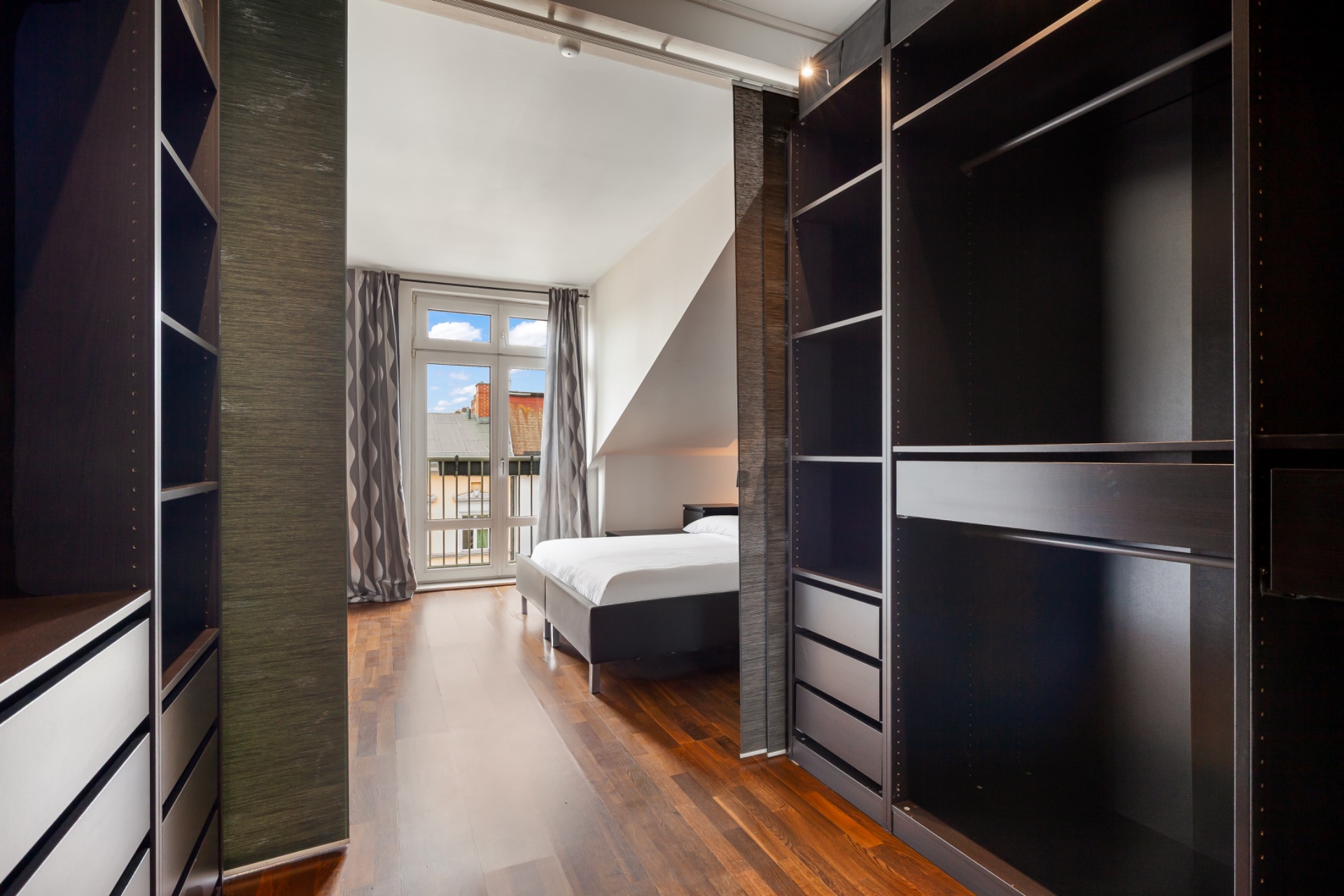 Schlafzimmer mit Holzboden, Doppelbett, großen Fenstern und offenen Regalen im Vordergrund.