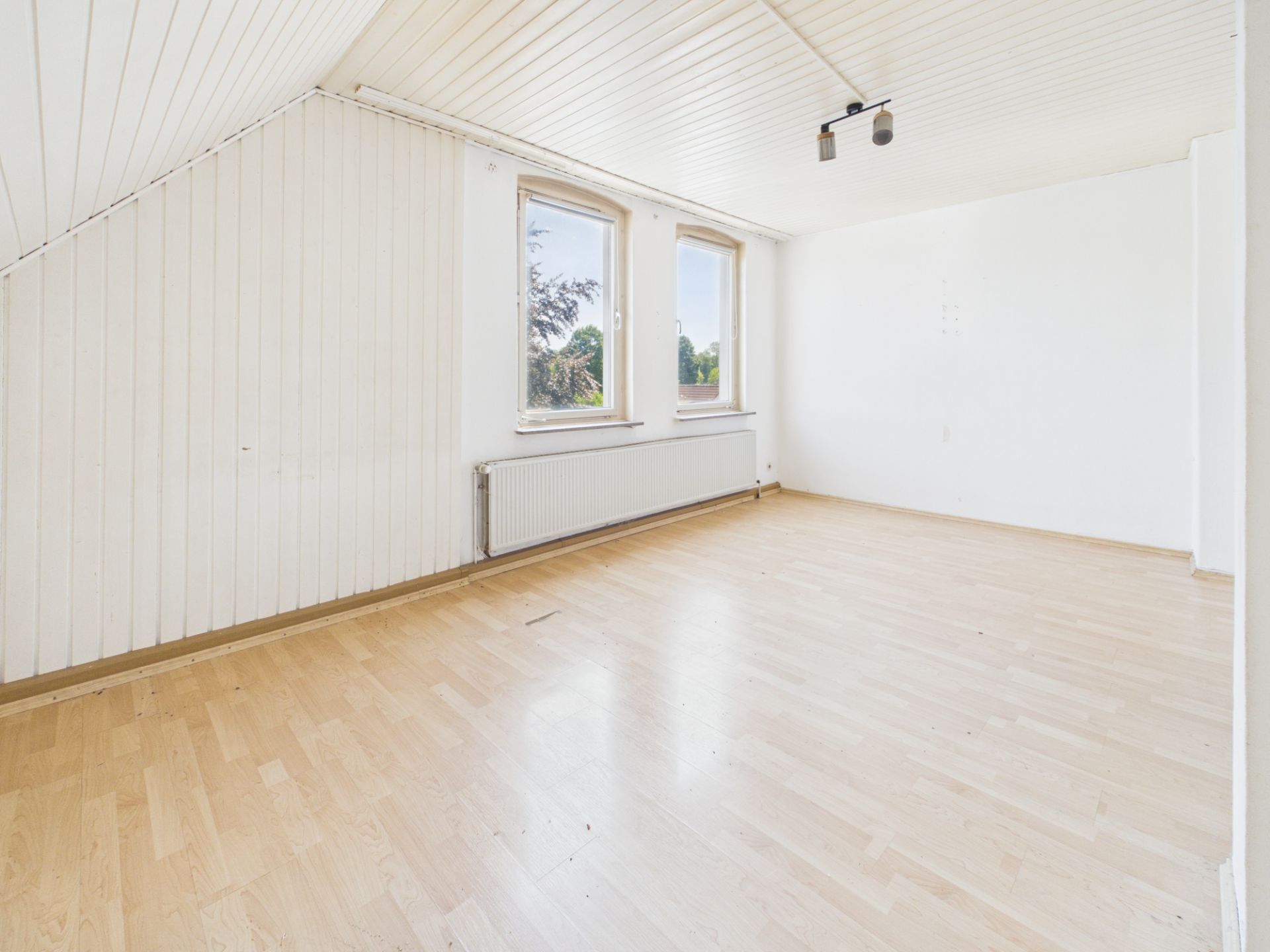 Helles, leeres Zimmer mit Holzfußboden, drei Fenstern und weißer Wandverkleidung.